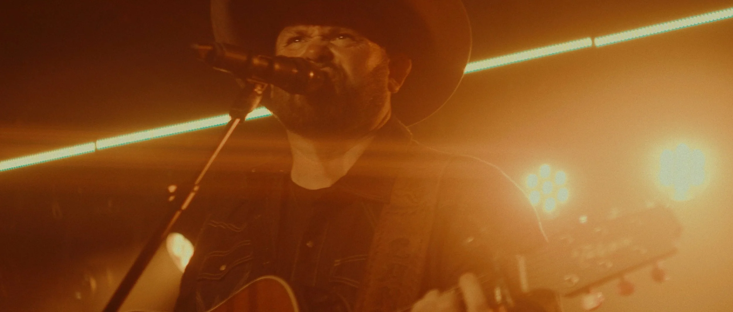 A man wearing a cowboy hat and dark clothing singing into a microphone while playing guitar on stage with vibrant orange and yellow lighting.
