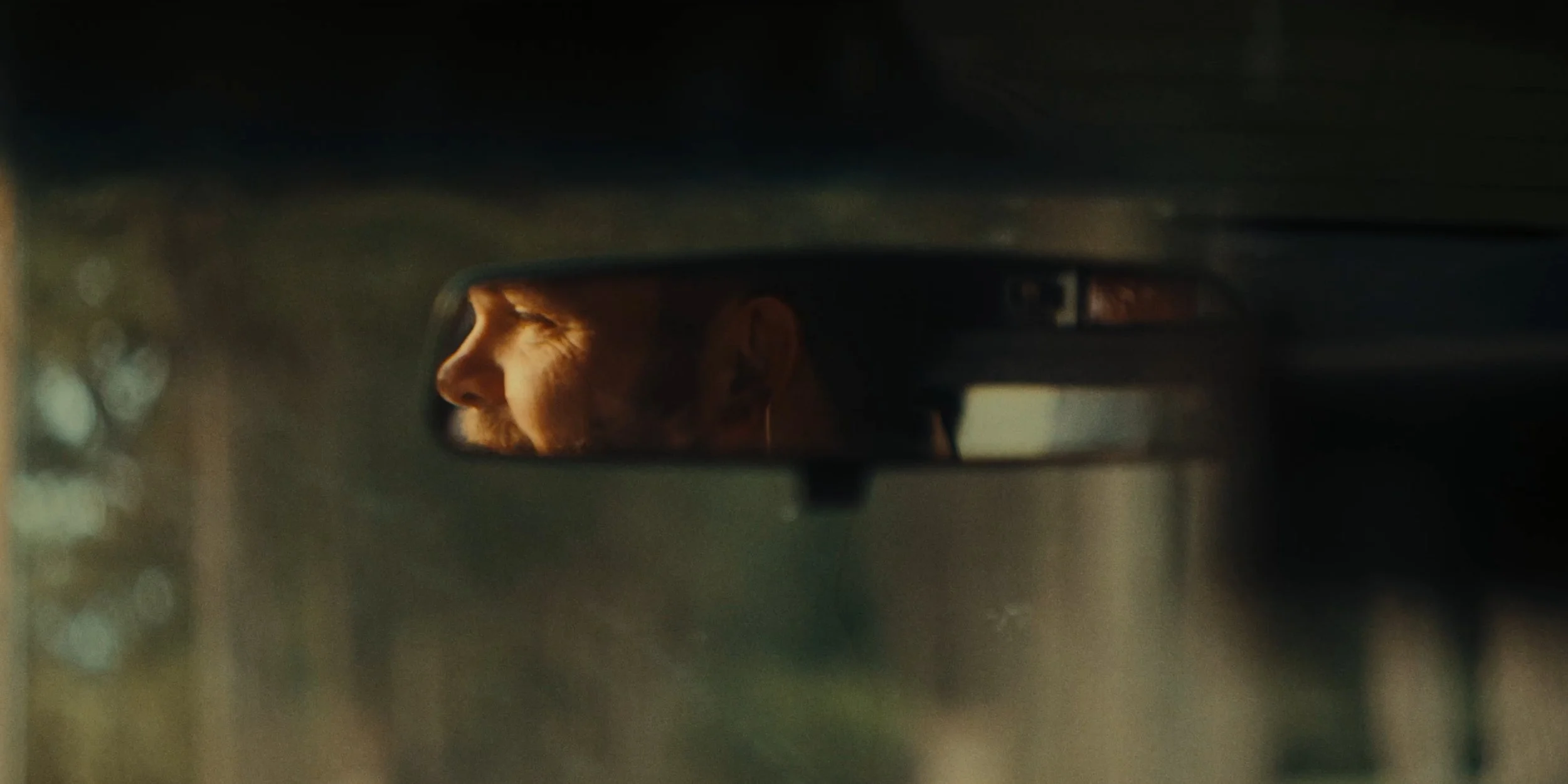 A person's profile reflected in a rearview mirror, with sunlight on their face and eyelid closed, sitting inside a vehicle.