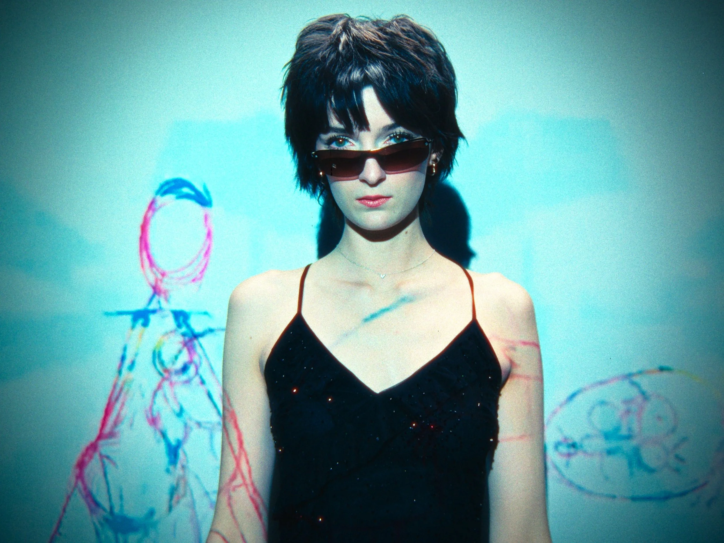 A woman with short dark hair wearing sunglasses and a black dress standing in front of a wall with colorful abstract drawings.