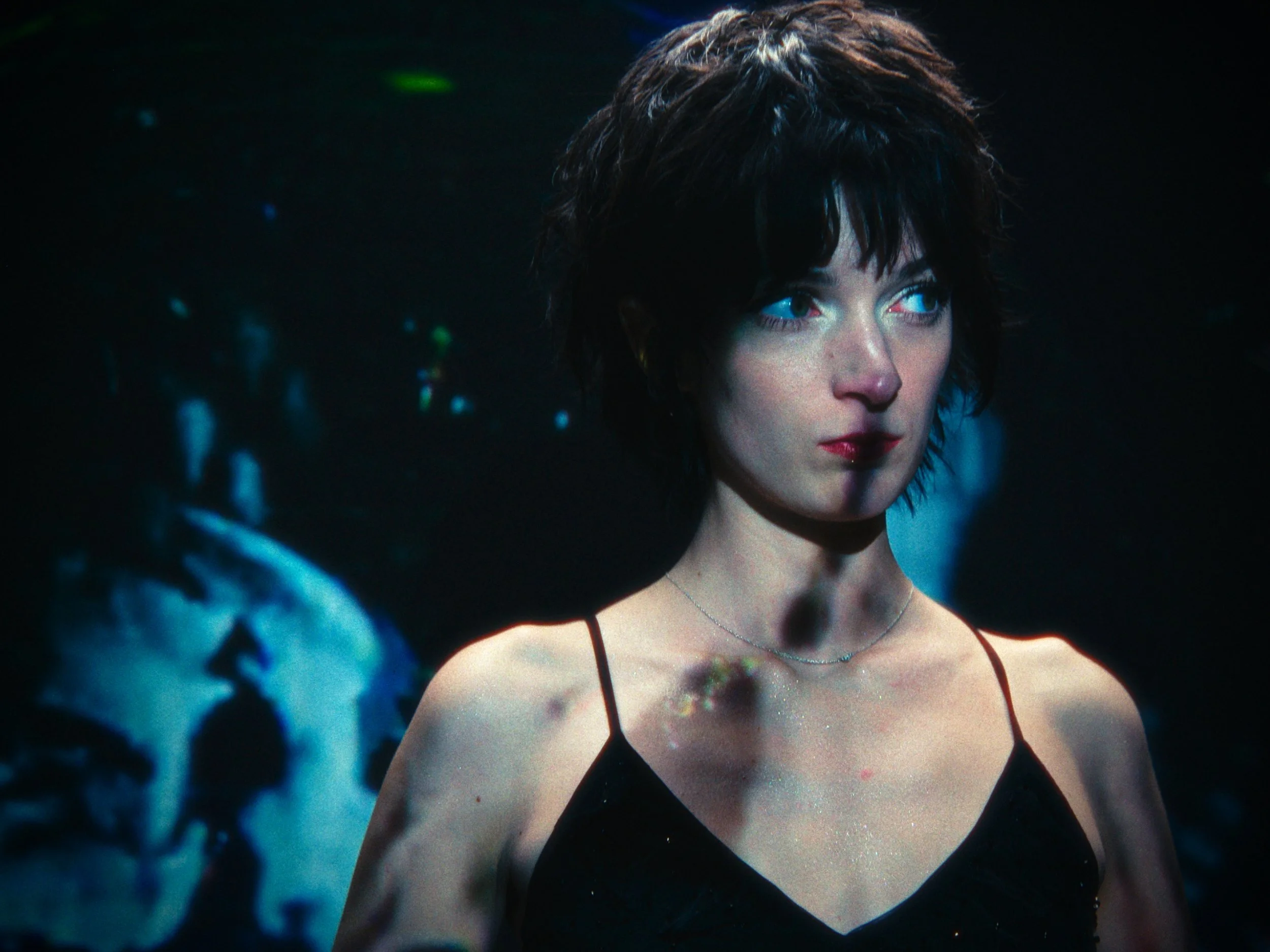 A woman with short dark hair and blue eyes, wearing a black spaghetti strap top, standing in a dark environment with faint blue and green lights. She has a serious expression and is looking to her right.