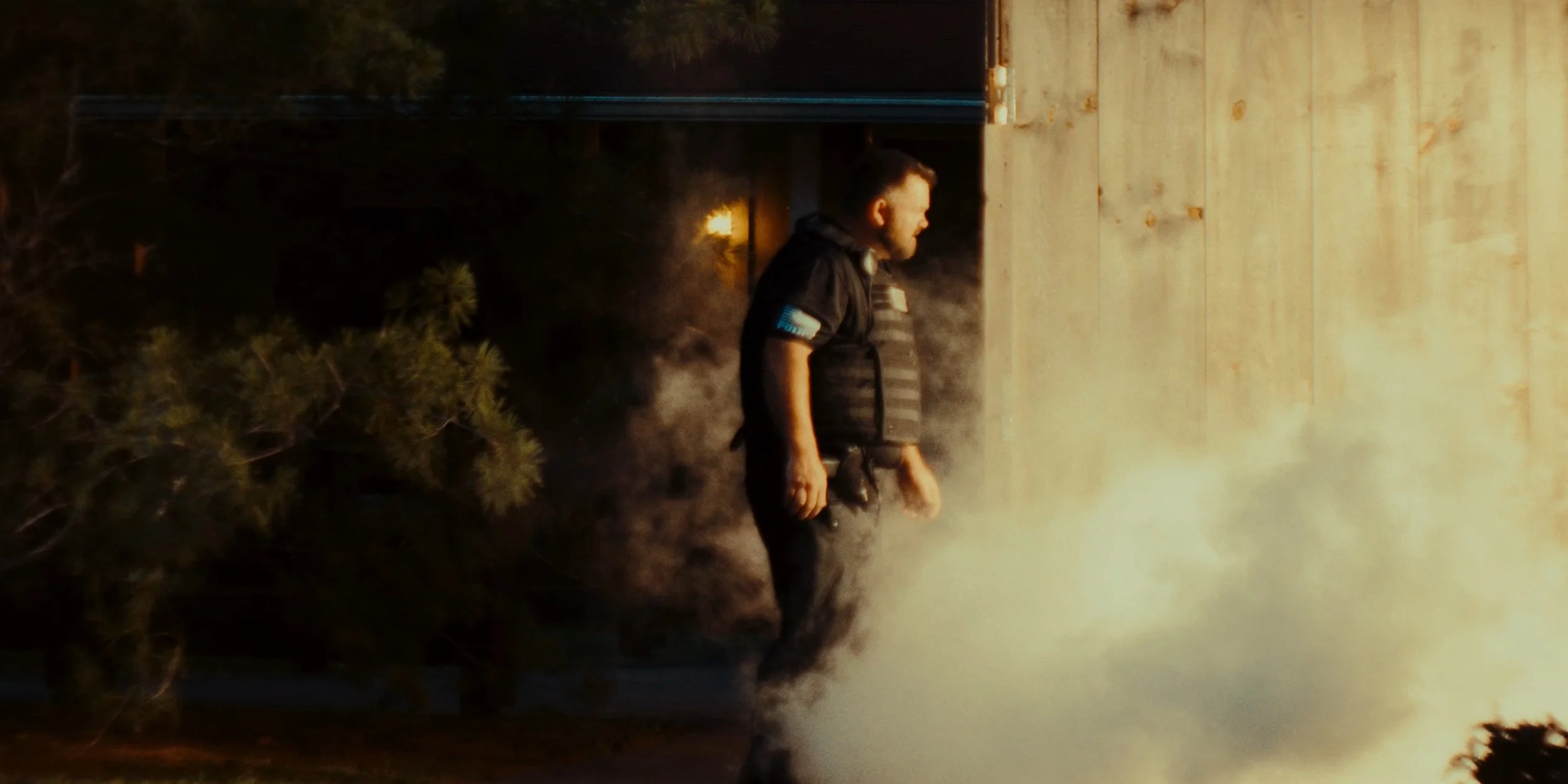 A person wearing a black vest standing in front of a fire with thick smoke, near a wooden wall.