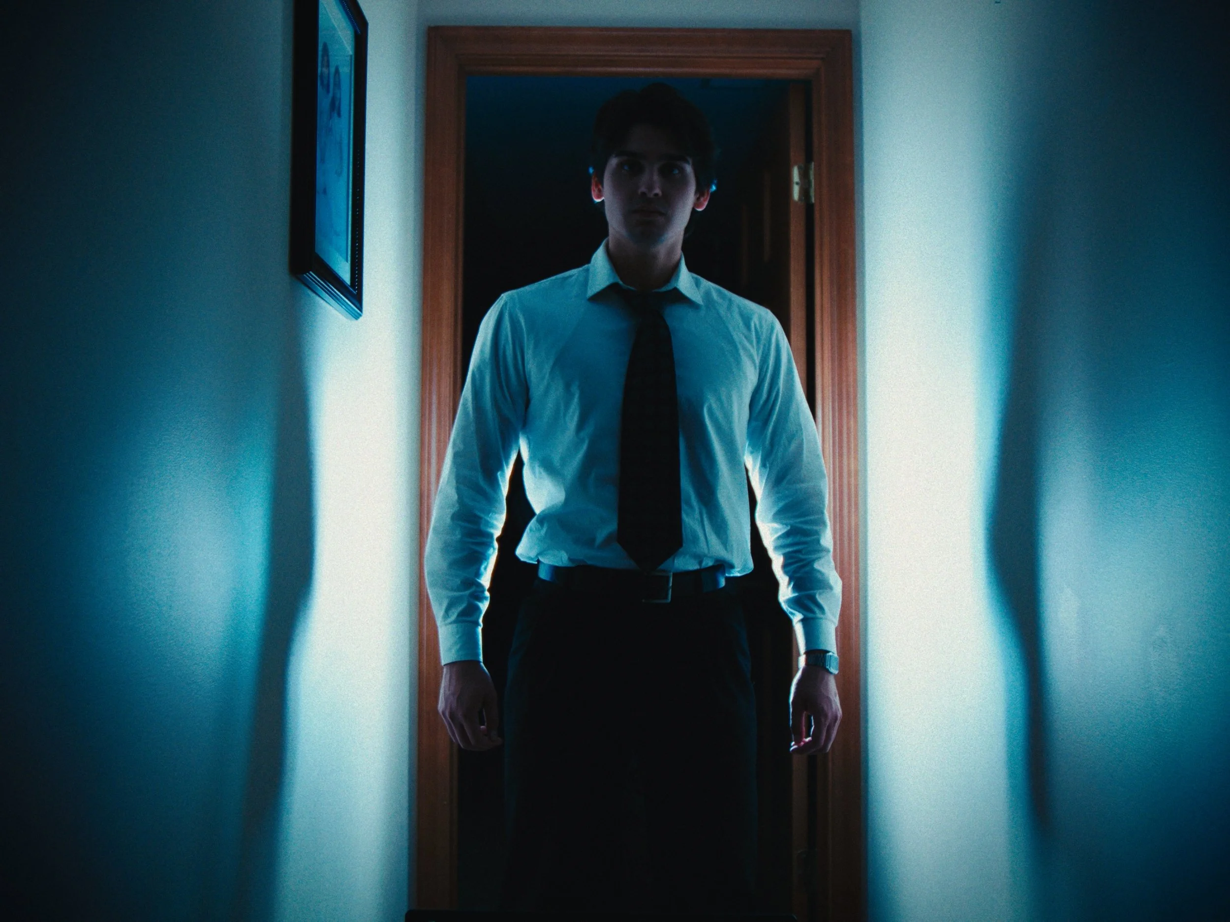 A young man in business attire, including a white shirt and dark tie, stands in a dimly lit corridor, facing forward with a serious expression, with light illuminating from behind.