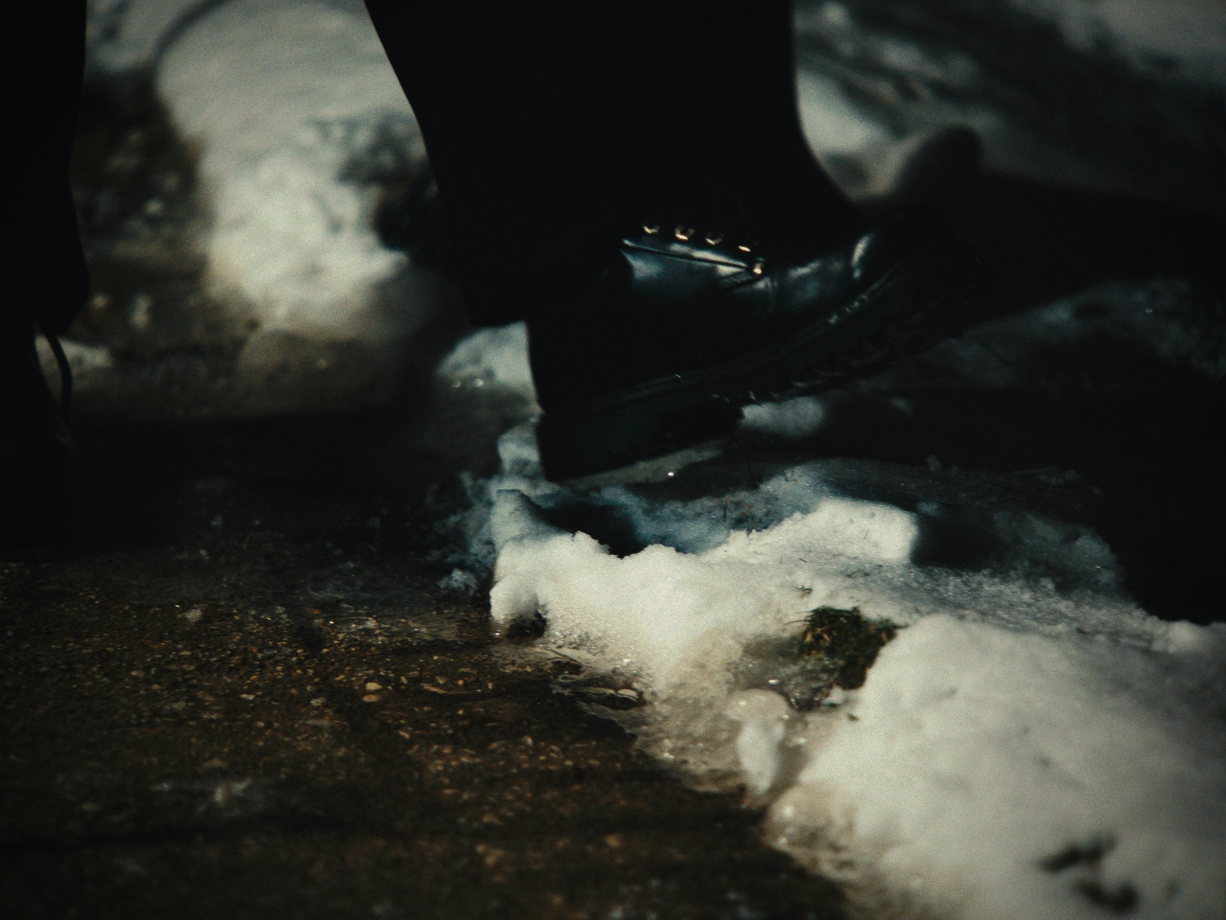Close-up of black boots stepping on snow and ice on a wet ground.