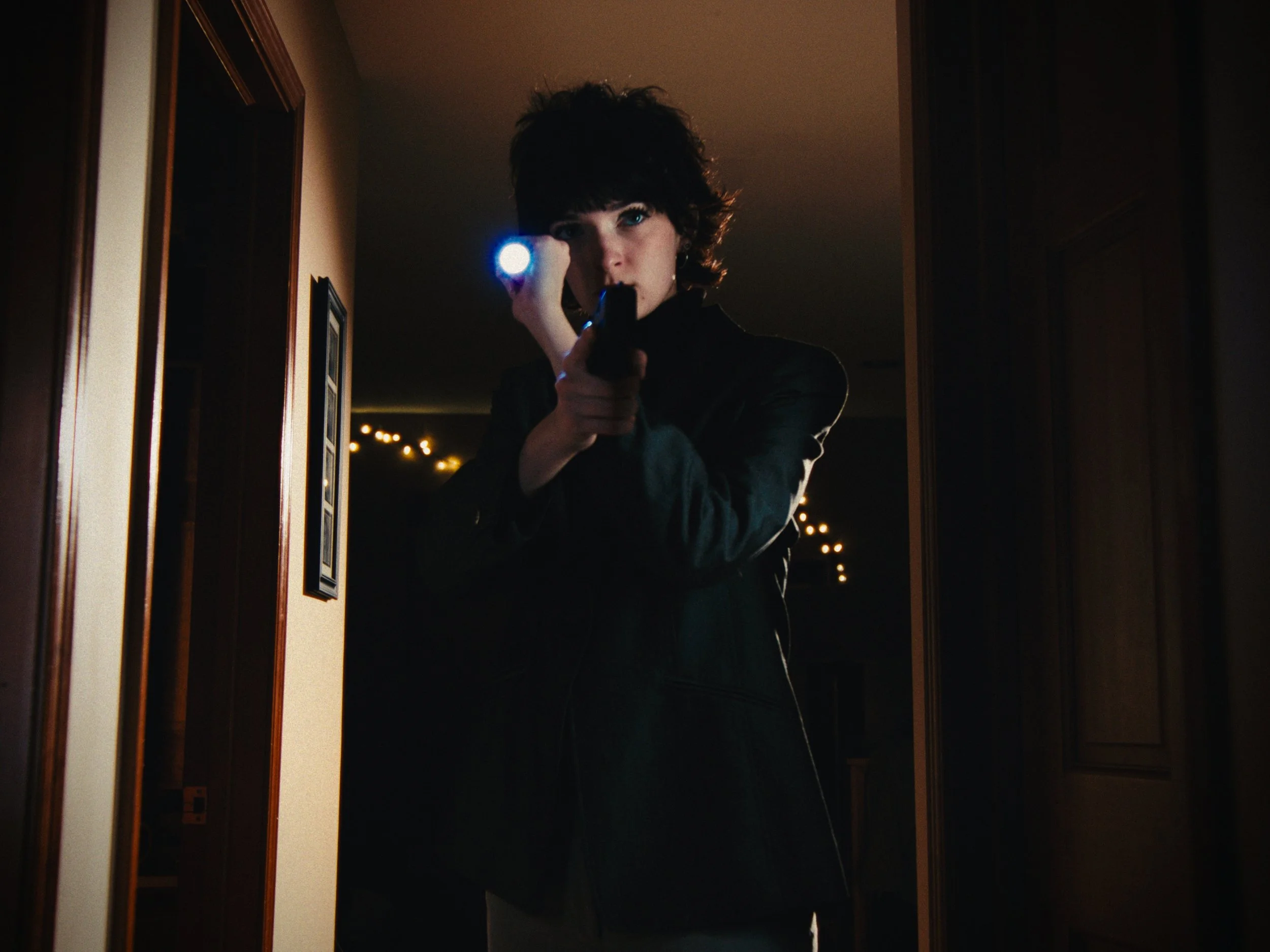 Person with short curly hair pointing a flashlight towards a mirror, revealing a gun in their hand, in a dimly lit room.