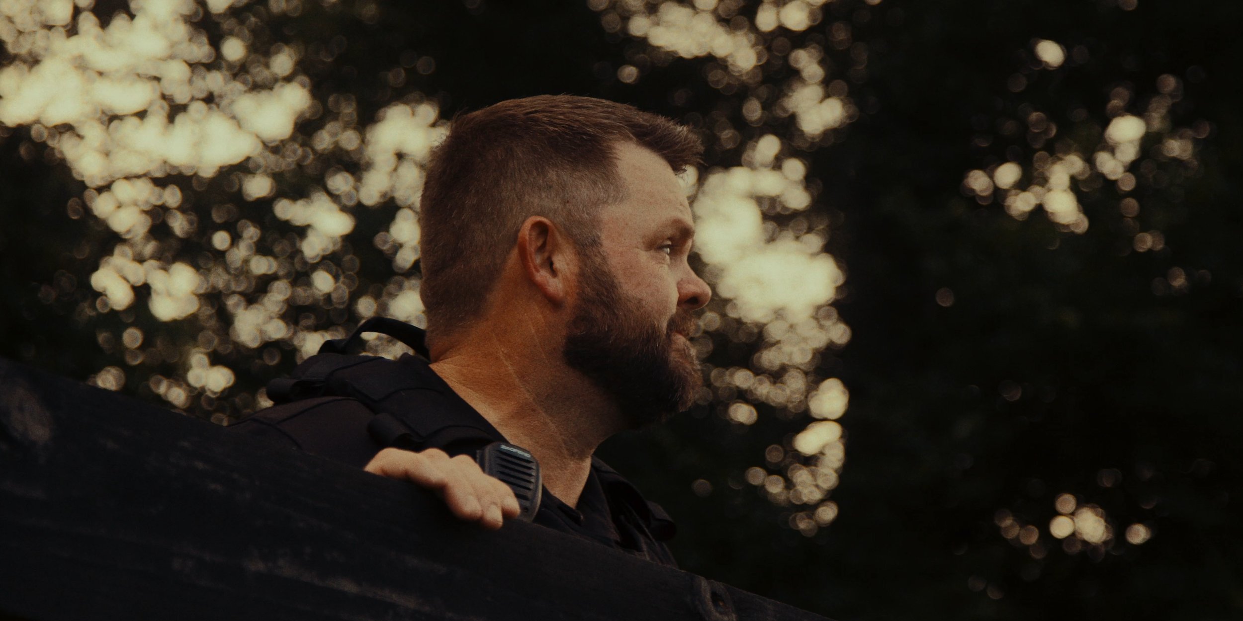 A man with a beard and short hair, wearing a dark uniform and a radio, looking to the side, amid blurred trees and bright sunlight in the background.