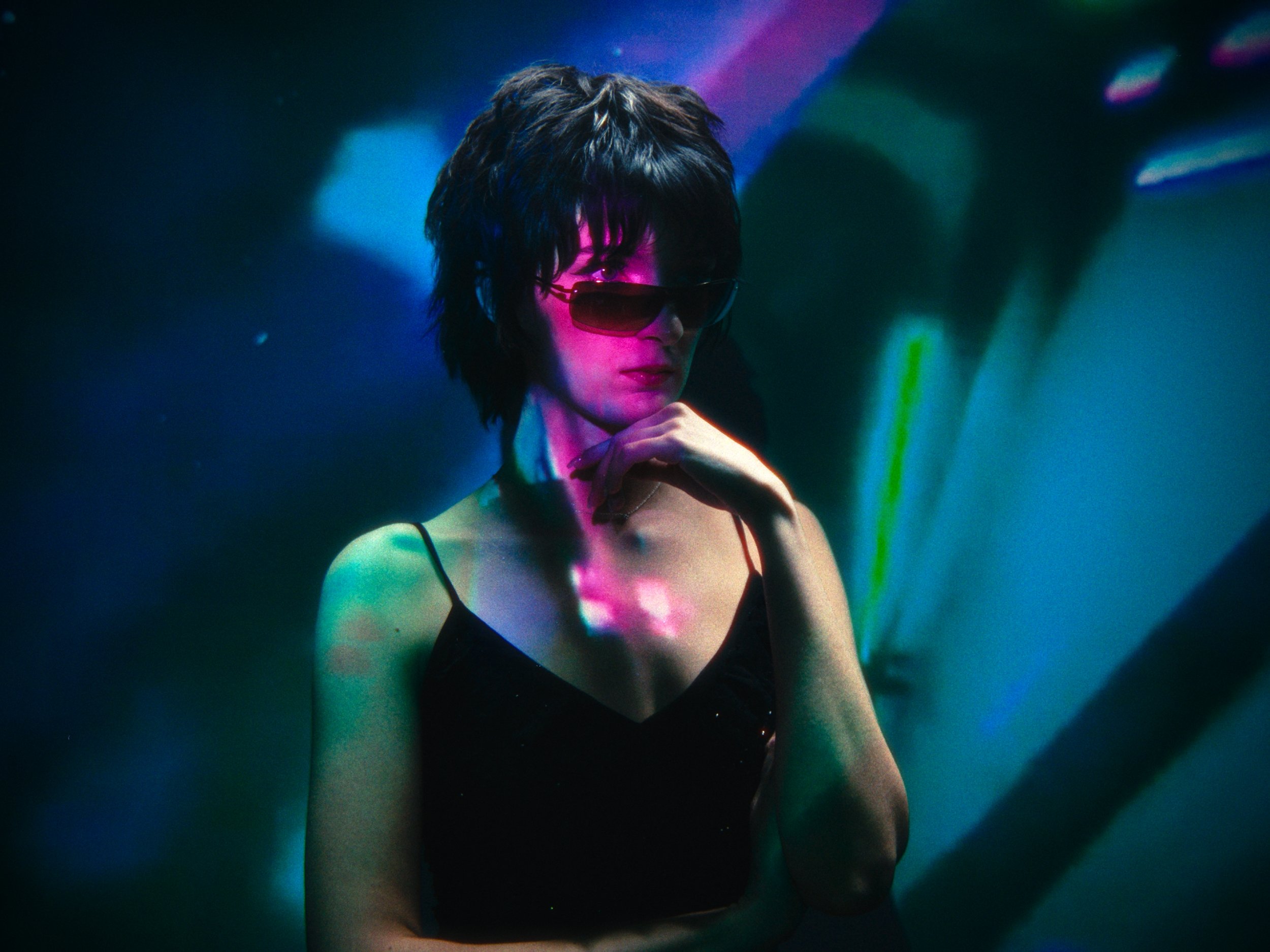 A woman with short black hair wearing sunglasses and a black sleeveless top, illuminated by colorful lights in a dark room.