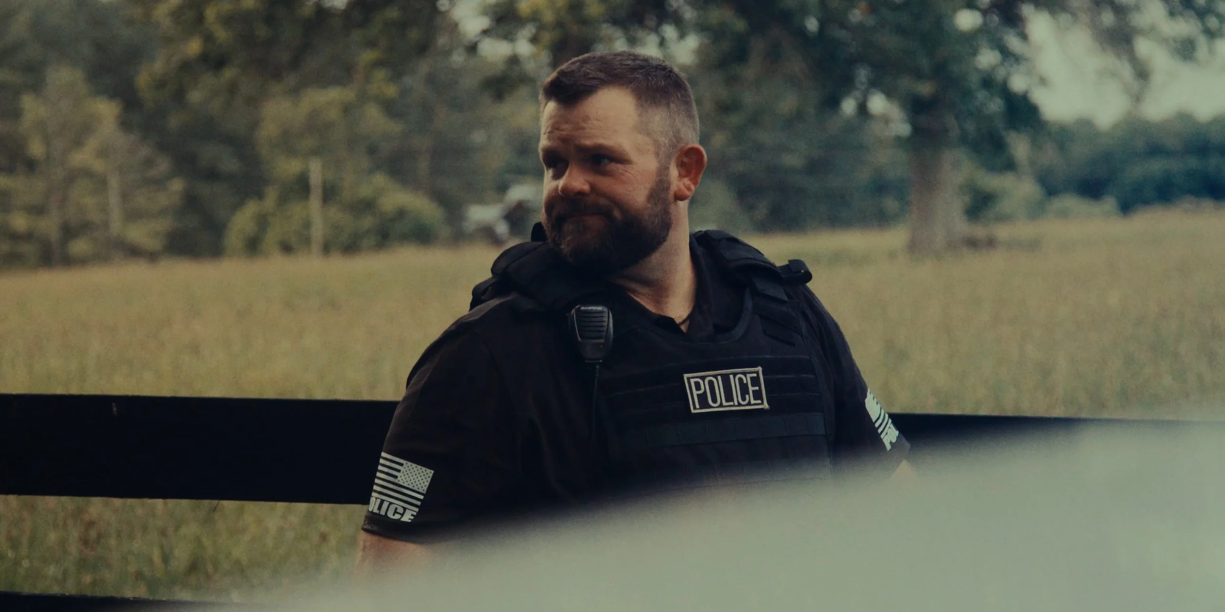 A male police officer with a beard and short hair, wearing a black tactical vest with a police badge, standing outdoors in a grassy area with trees in the background.