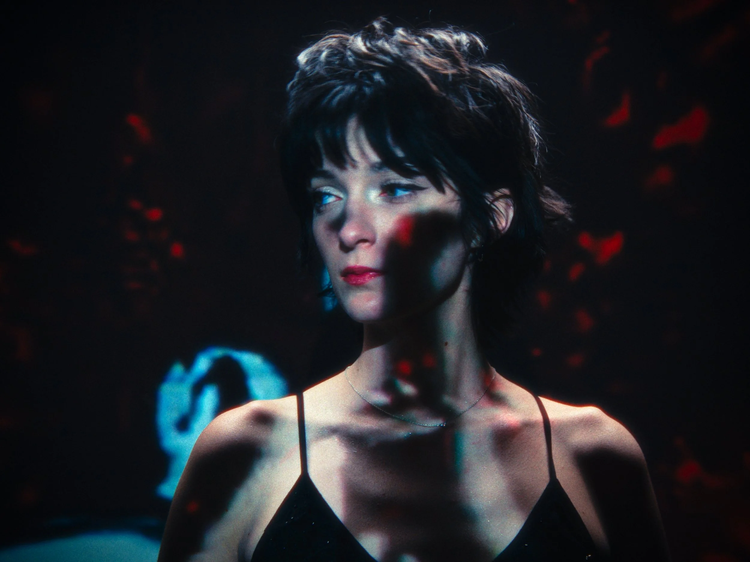 A woman with short dark hair and blue eyes looking to her left, wearing a black spaghetti strap top, with red and blue reflections or lights on her face and background.