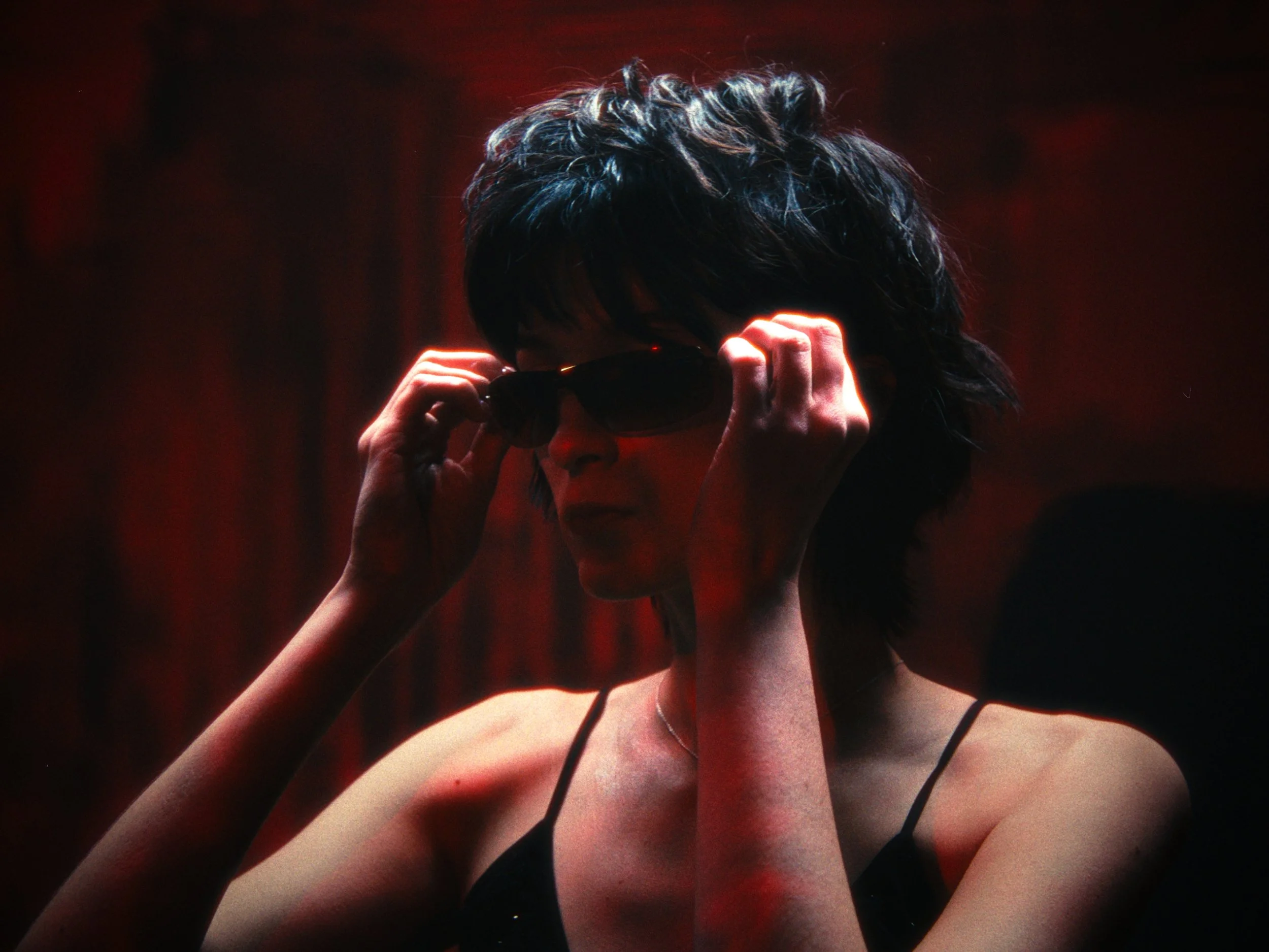 A woman adjusting sunglasses in a dimly lit setting with red lighting, black tank top, and short dark hair.