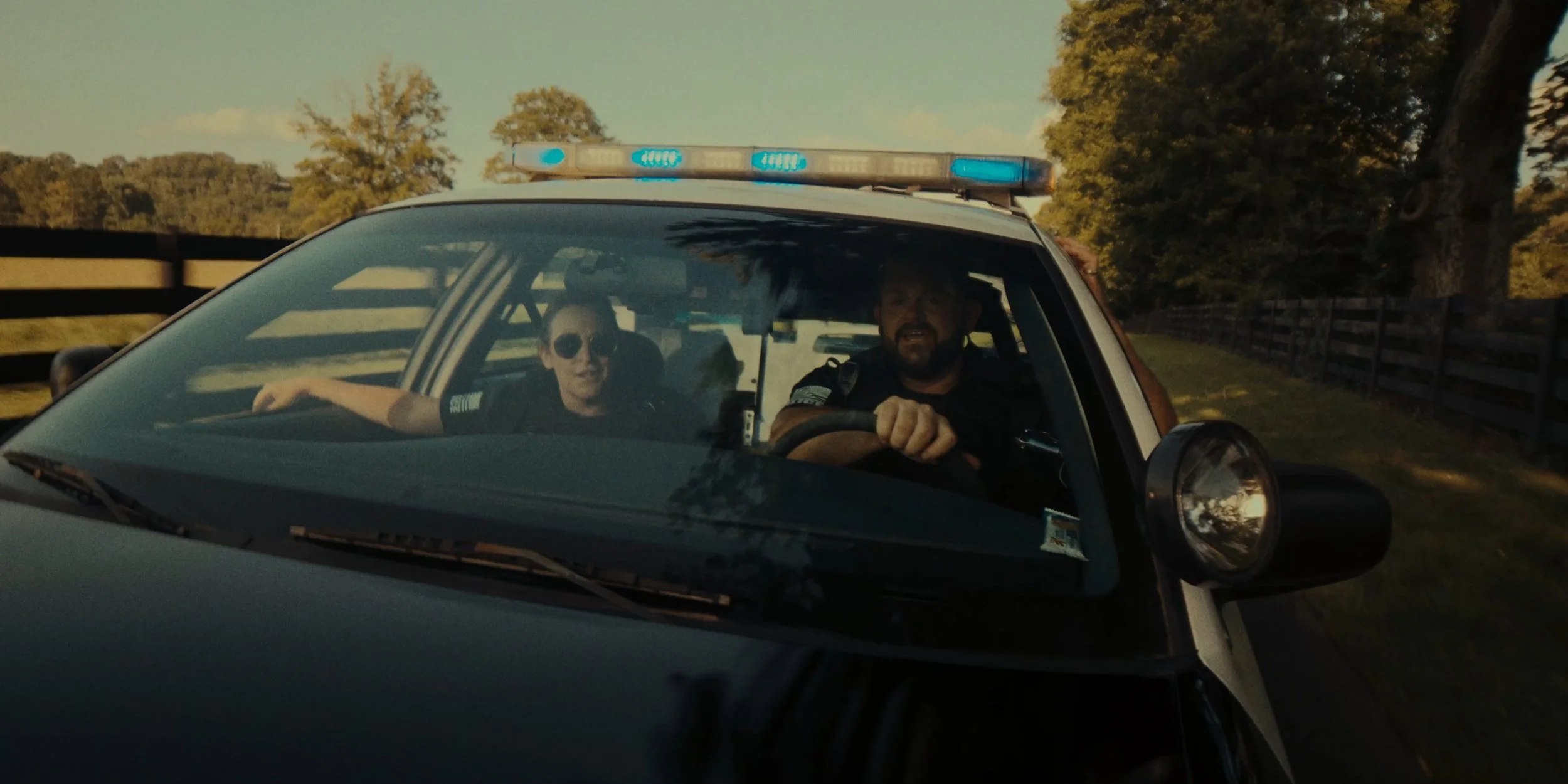 A police car with flashing blue lights on the roof, containing two officers inside, driving down a road with trees and a wooden fence in the background.