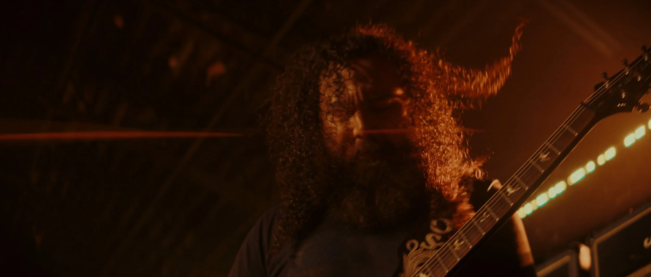 Man with long curly hair playing an electric guitar in a dark setting with warm lighting.