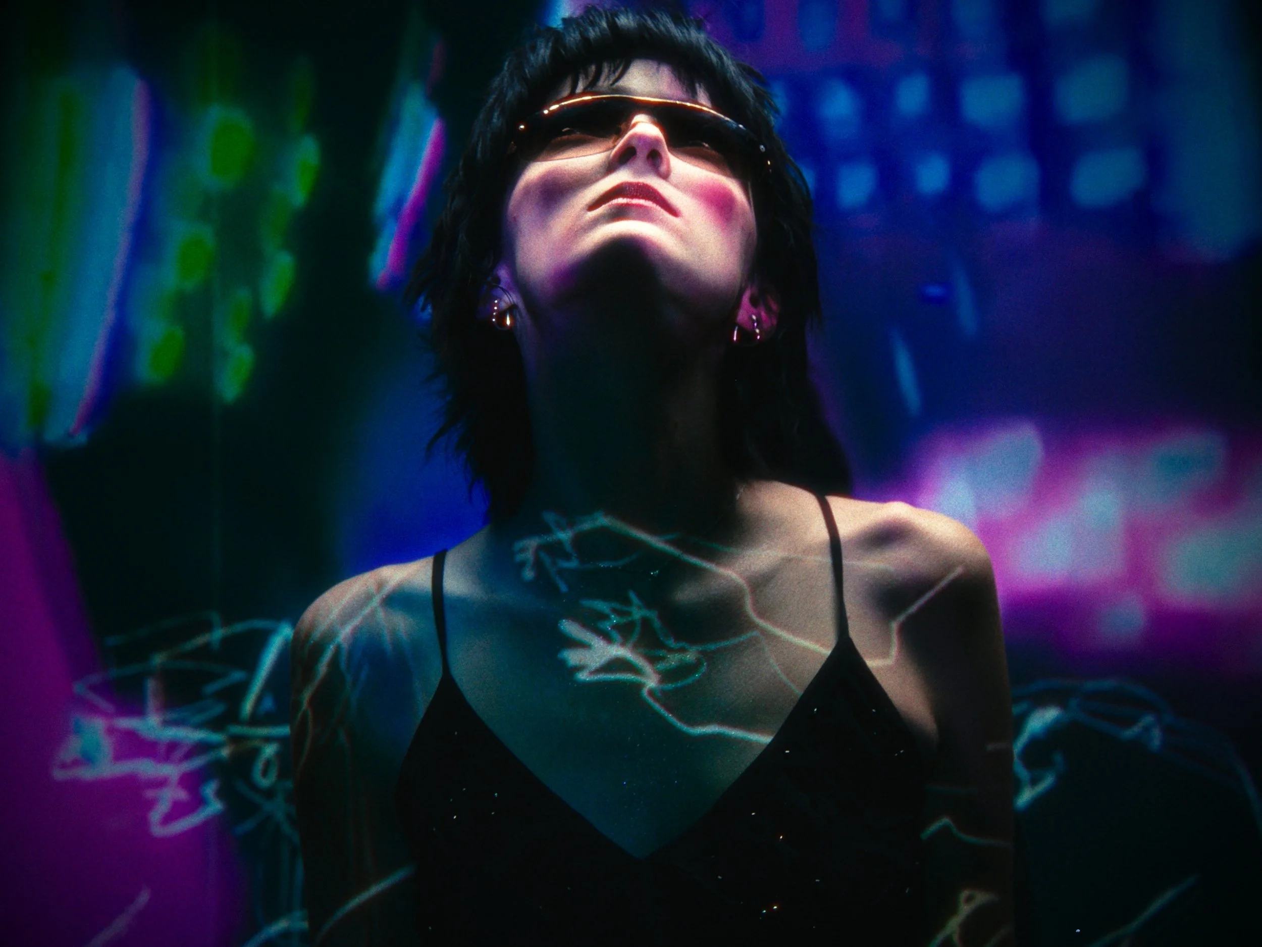 A woman with short dark hair, wearing sunglasses and earrings, is illuminated by neon lights with a dark background. She is wearing a black spaghetti strap top, and colorful abstract and lightning-like projections are visible on her chest and shoulde