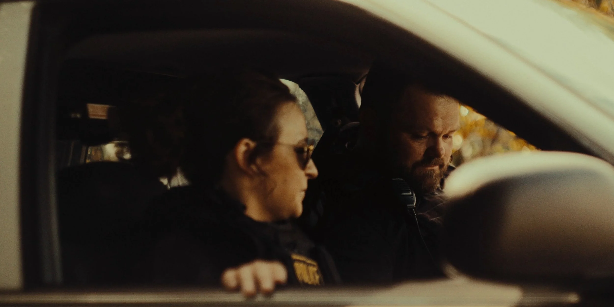 A woman and a man sitting in a car, both wearing police uniforms, with the woman wearing sunglasses, looking down, and the man looking down with focus.