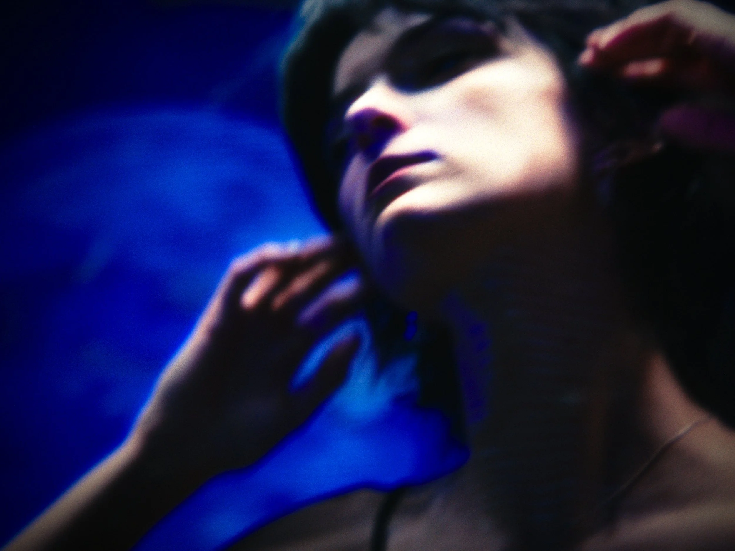 A woman with dark hair, partly covered by a hat, looking upwards with her hand near her ear in a dimly lit environment with blue and purple tones.