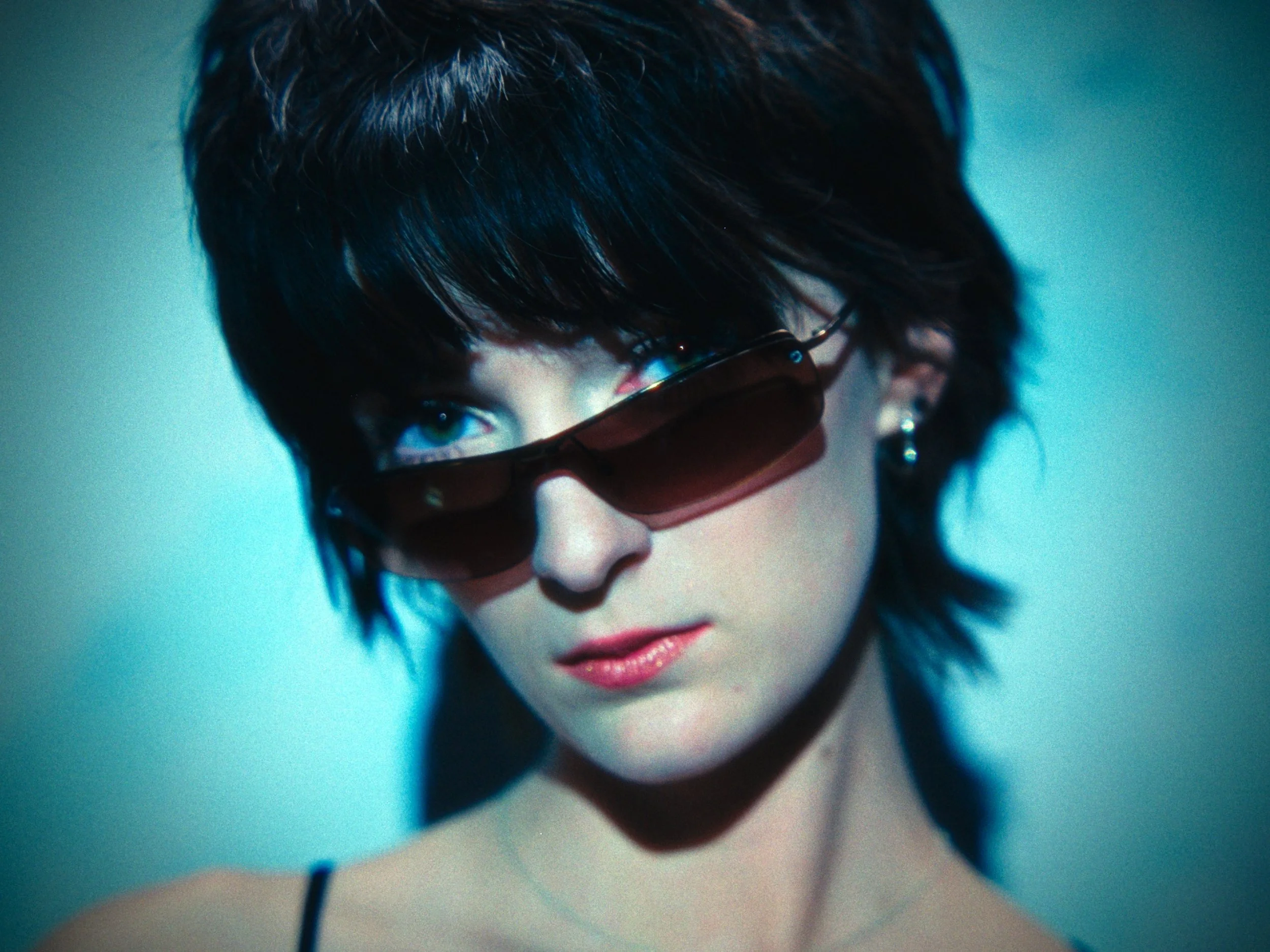 Close-up of a woman with short dark hair wearing brown sunglasses and a black strap top, with a confident expression.