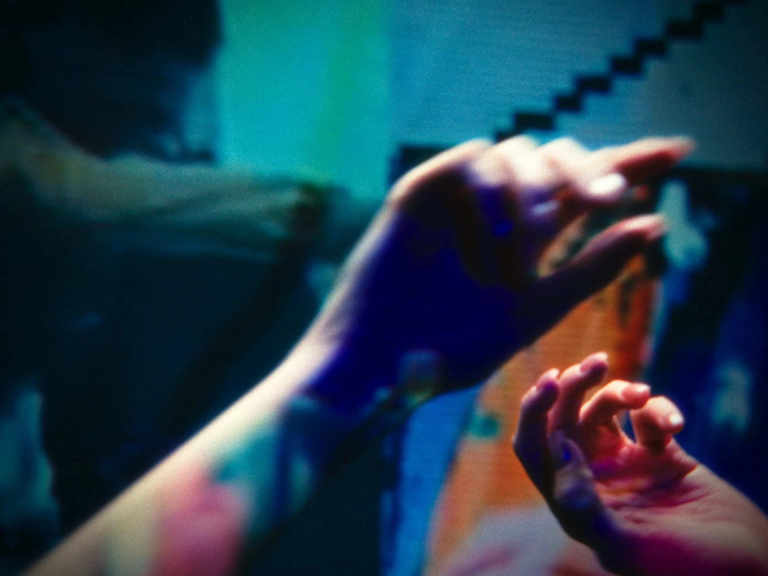 Close-up of hands with painted nails reaching towards a mirror, with a blurred background and colorful lighting.