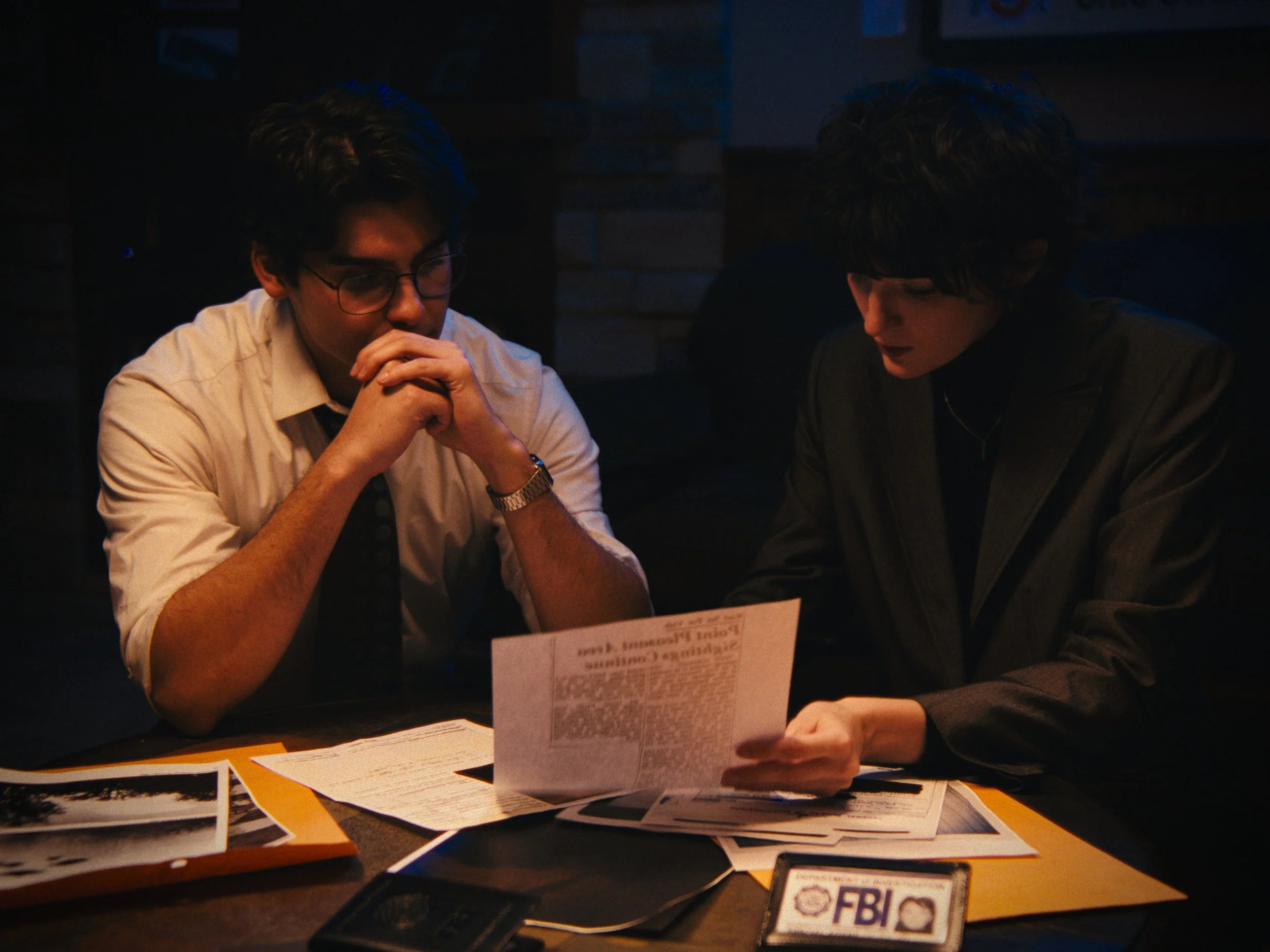 Two people sitting at a table reviewing documents and photos, with an FBI badge prominently displayed on the table, in a dimly lit setting.