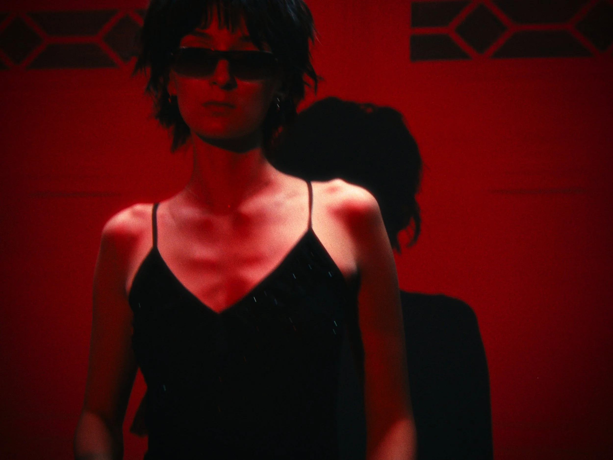 A woman with dark hair wearing sunglasses and a black top with spaghetti straps stands in front of a red background.
