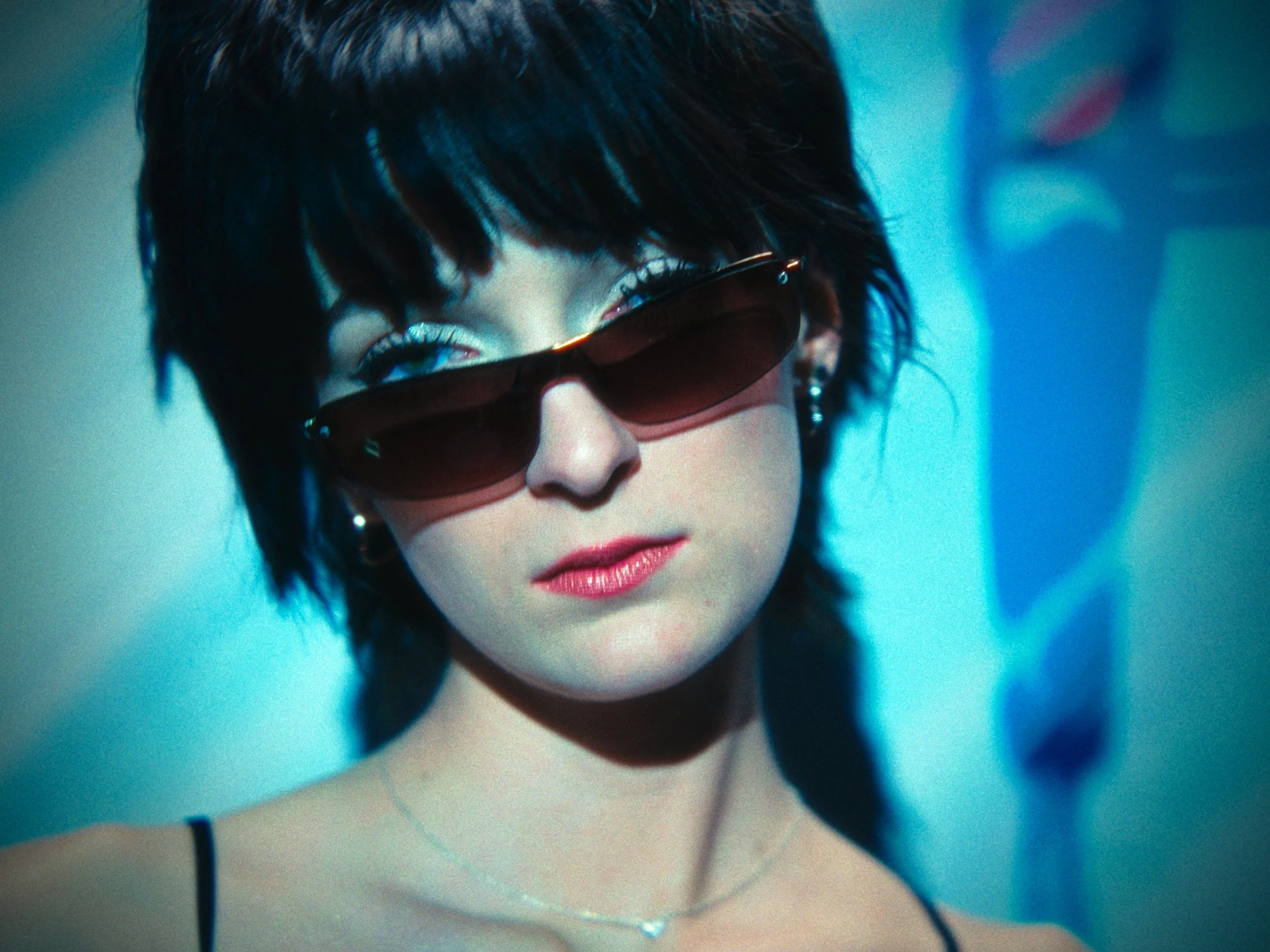 Close-up of a woman with short dark hair wearing sunglasses and a necklace, with makeup and a strappy top.