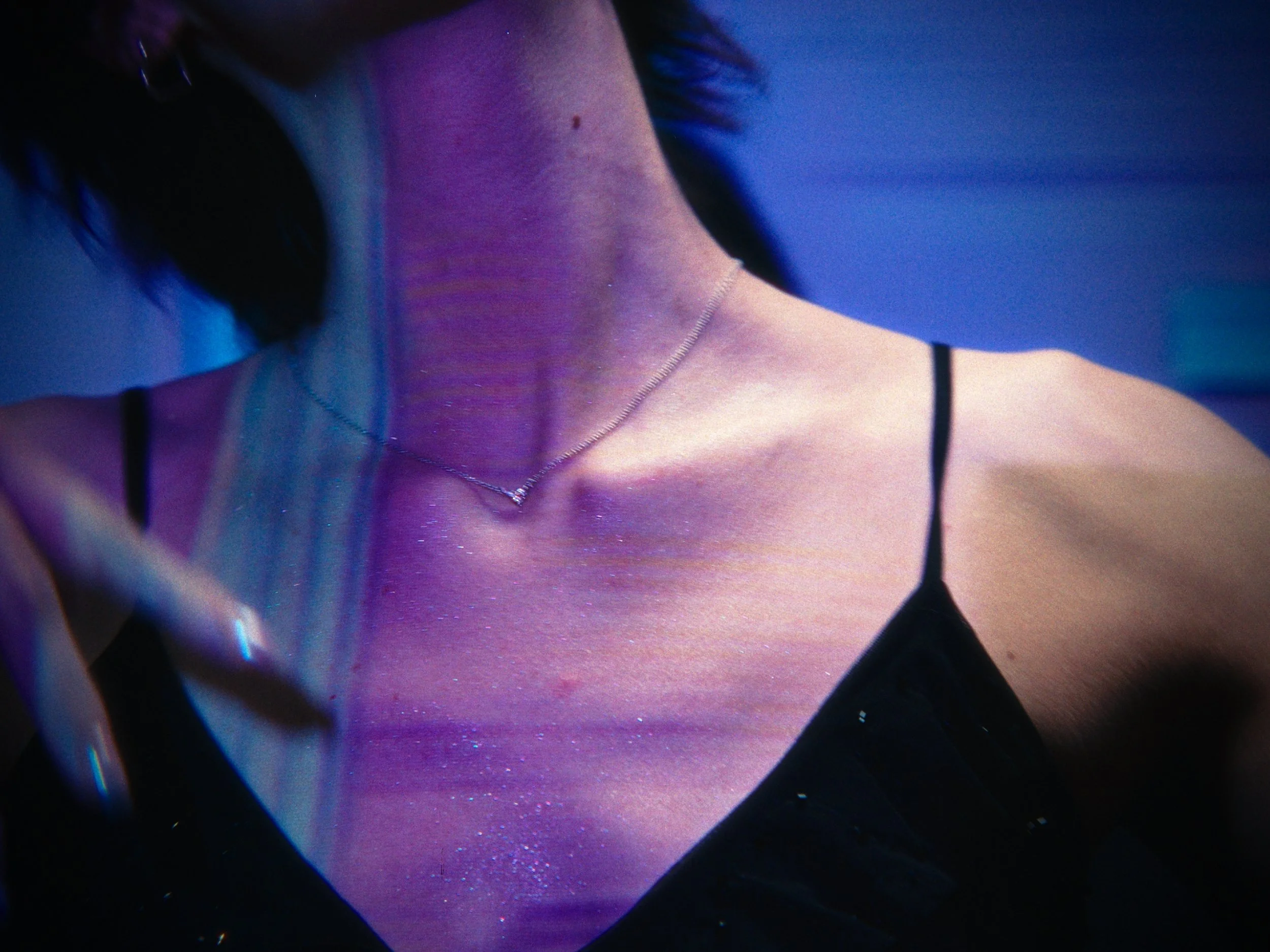 Close-up of a woman's neck and shoulder under colorful, shiny lighting, wearing a delicate necklace and a black spaghetti strap top.
