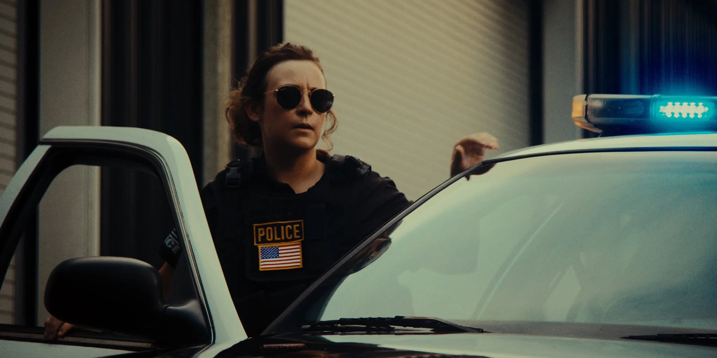 Female police officer with sunglasses standing beside a police vehicle with blue lights on, in an urban setting.