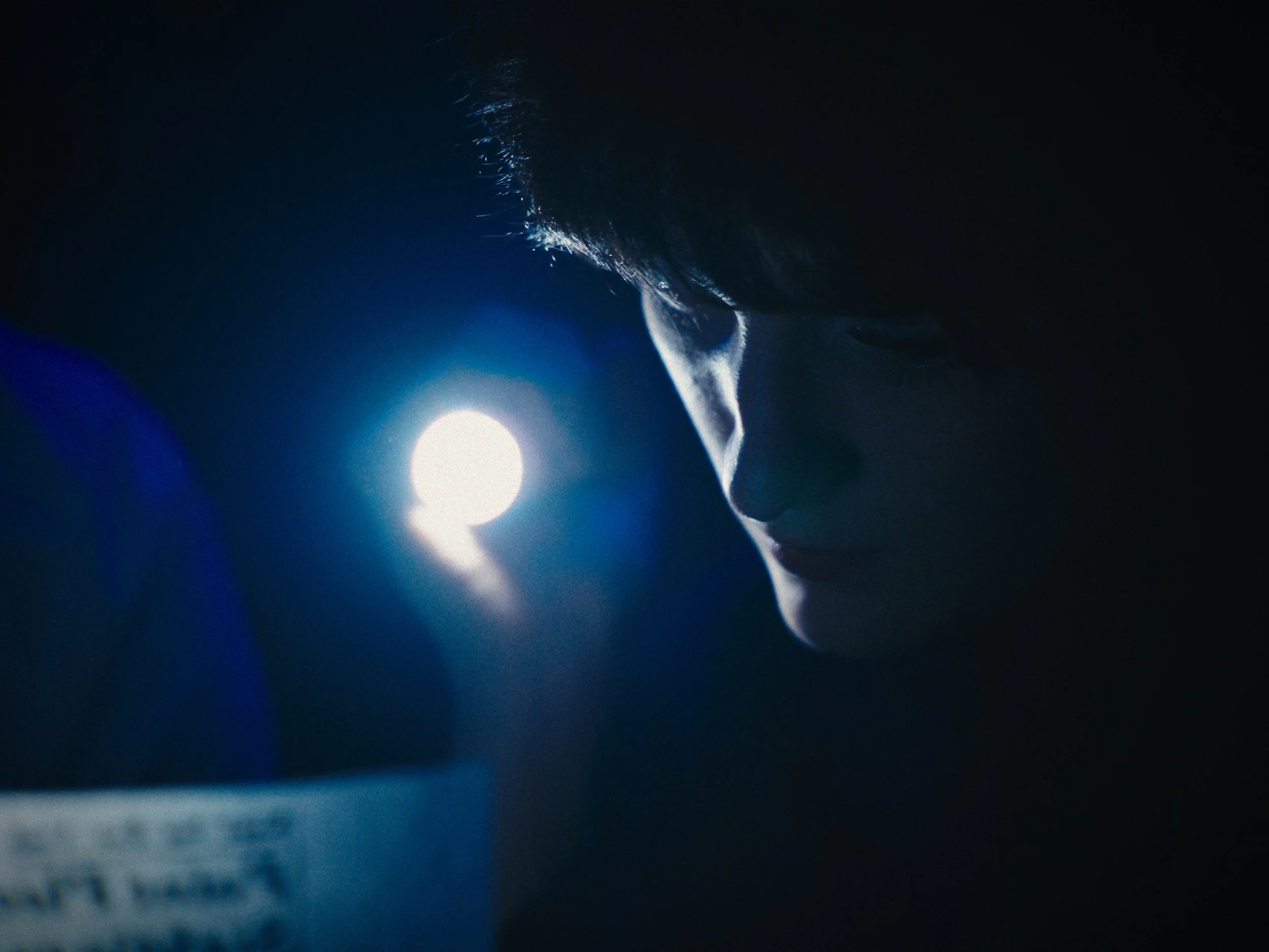 A person with dark hair looking down in a dimly lit environment with a bright light source nearby.