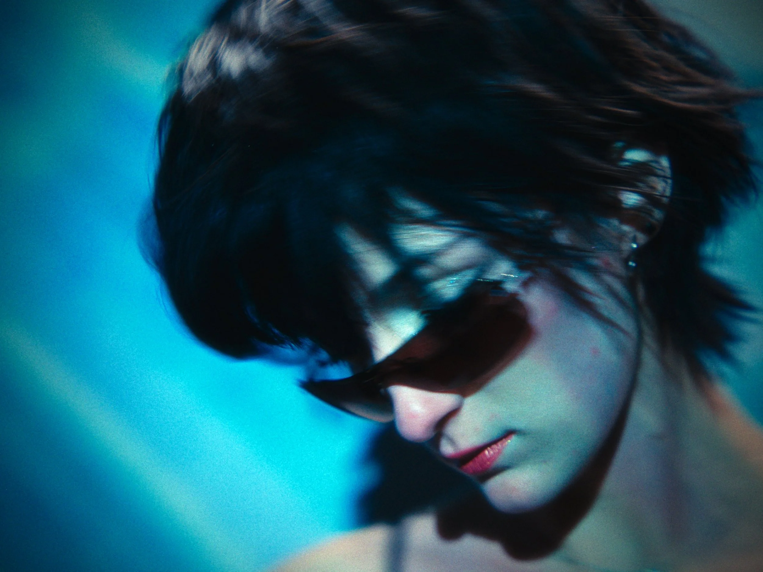 A woman with dark, wet hair wearing black sunglasses, looking down against a blue background.