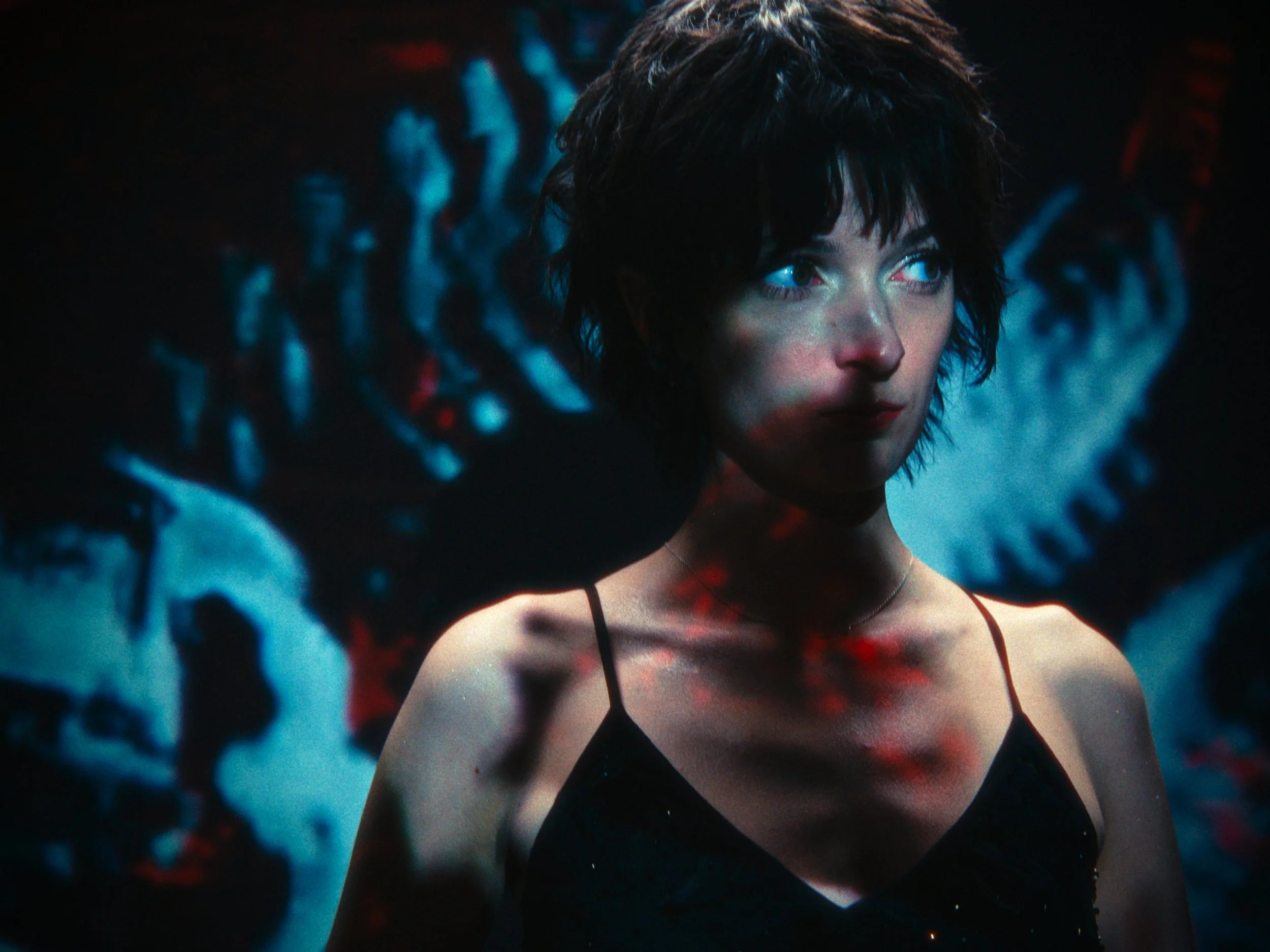 A woman with short dark hair, wearing a black spaghetti strap top, looking to the side with a serious expression, surrounded by a dark scene with blue and red abstract patterns.