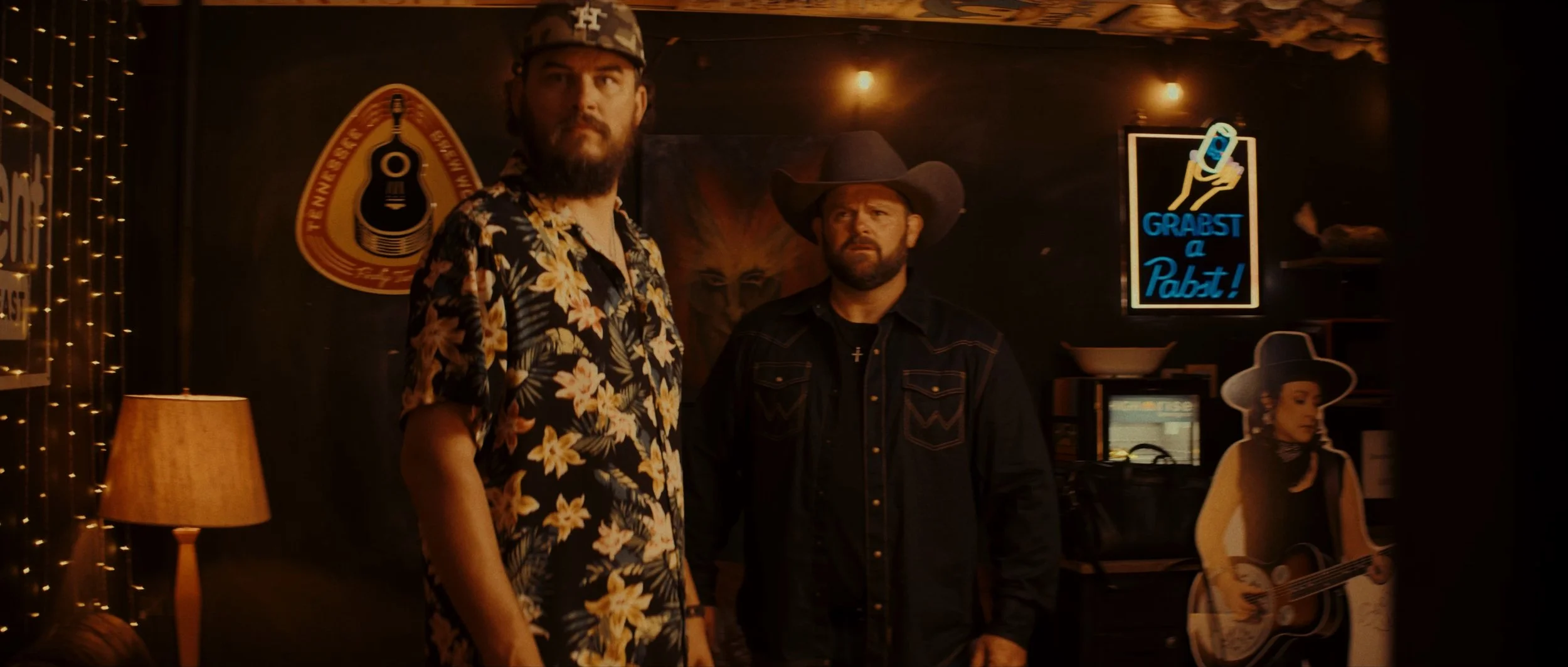 Two men standing indoors near wall decorations, one wearing a floral shirt and the other a cowboy hat. There are neon signs, including one that says 'GRABST a Pabst!', and a cardboard cutout of a woman with a guitar, all in a dimly lit environment.