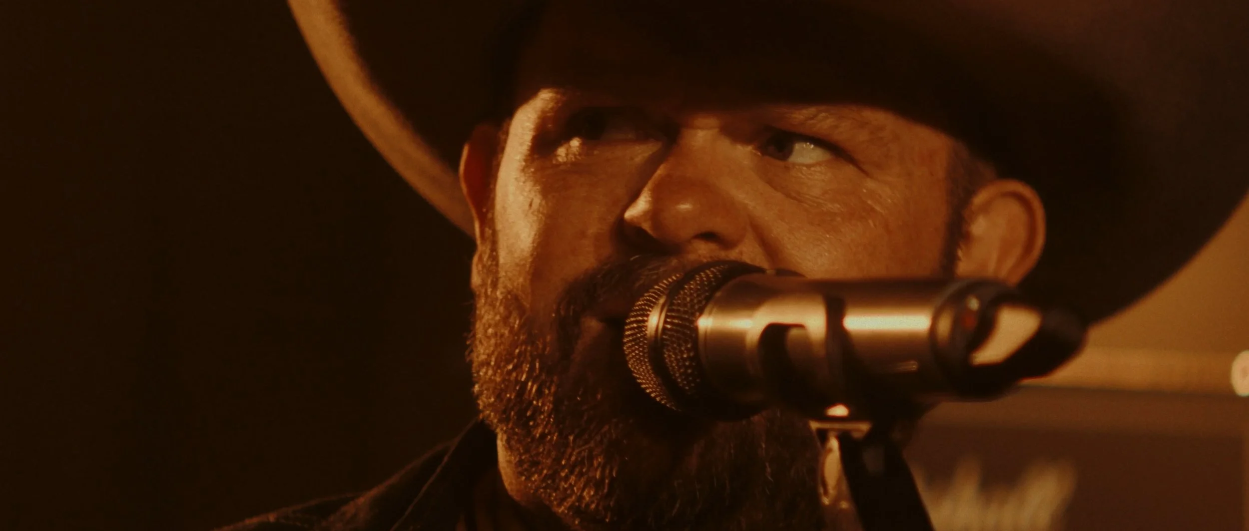Close-up of a man singing into a microphone, wearing a black cowboy hat, with a beard, in a dimly lit setting.