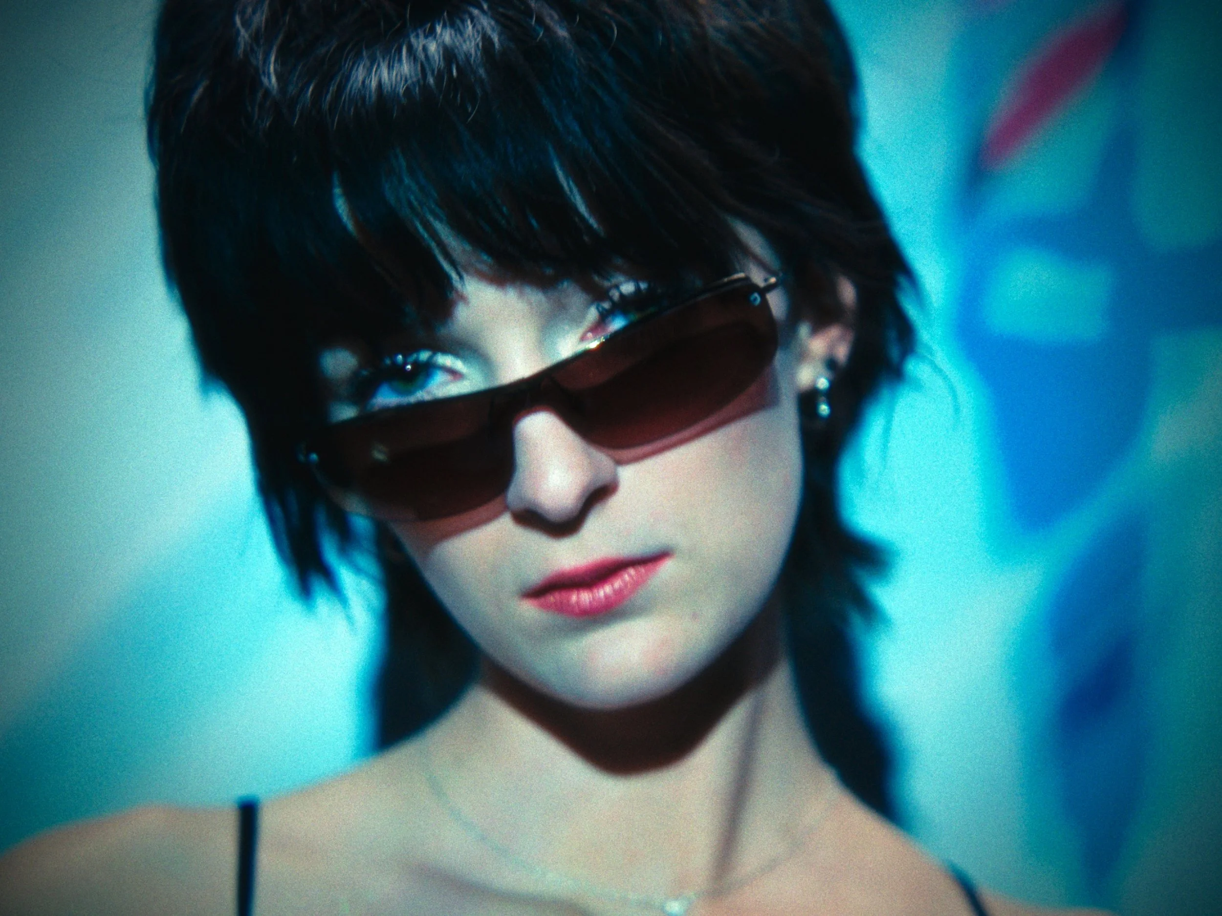 Close-up of a woman with short dark hair, wearing large sunglasses and earrings, looking confident with a blurred background.