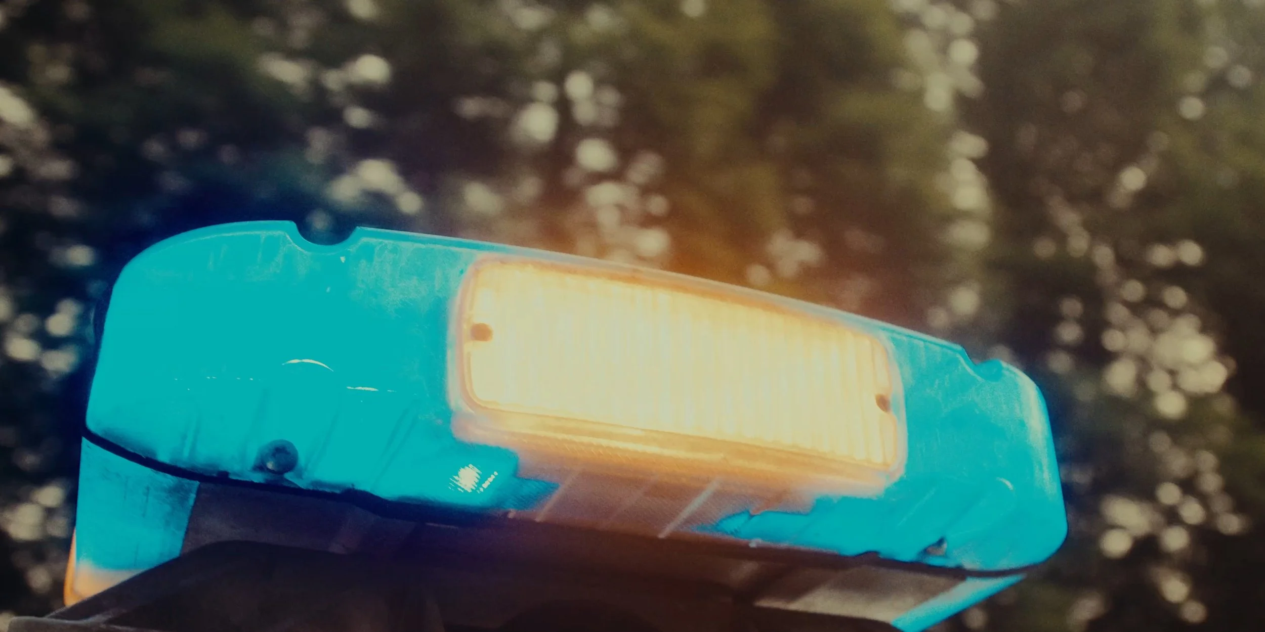 Close-up of a blue emergency or police light with yellow-orange lens, mounted on a vehicle, with blurred trees in the background.
