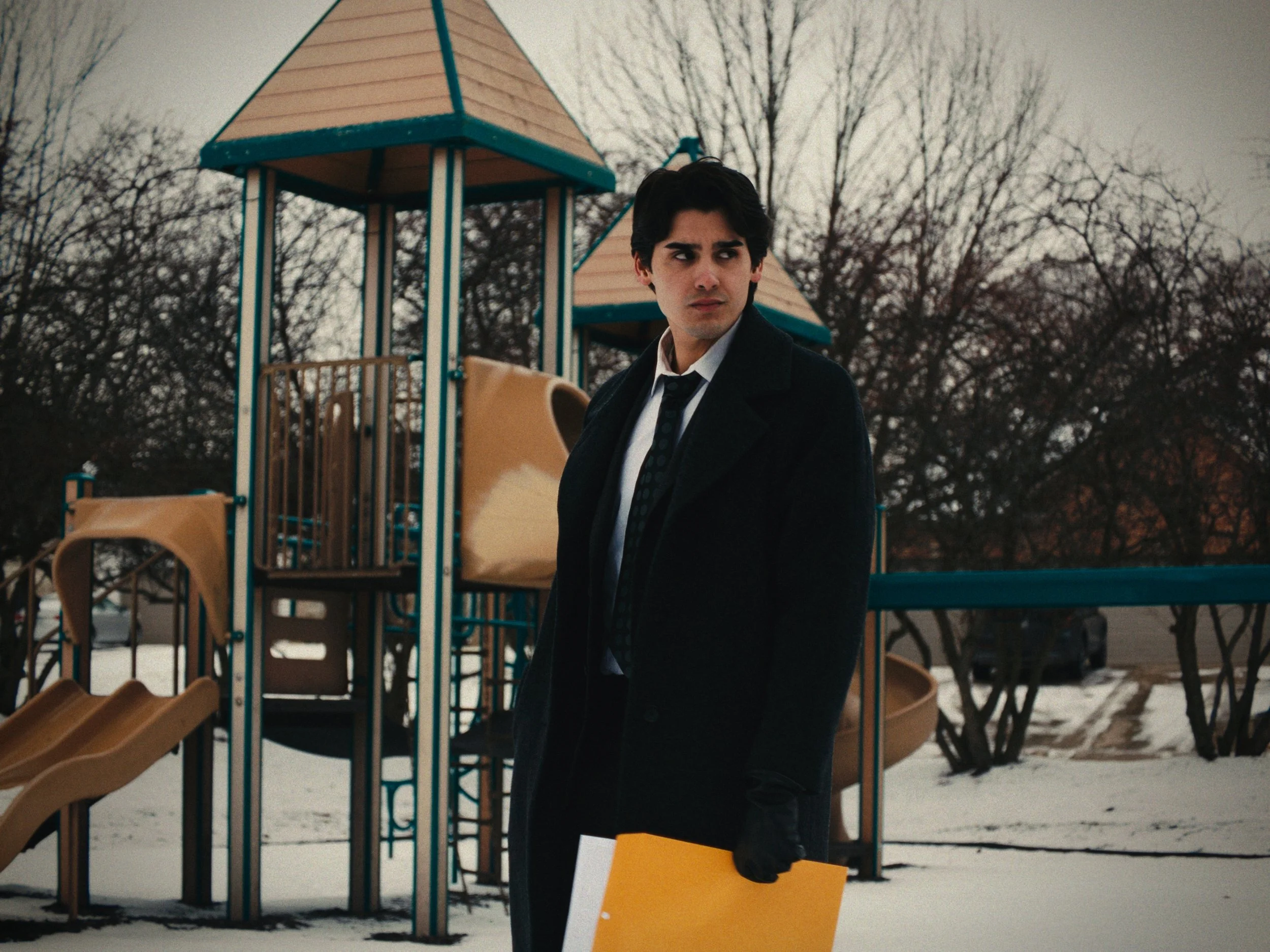 A young man in a black coat and gloves standing outdoors in front of a playground with snow on the ground, holding a yellow folder.