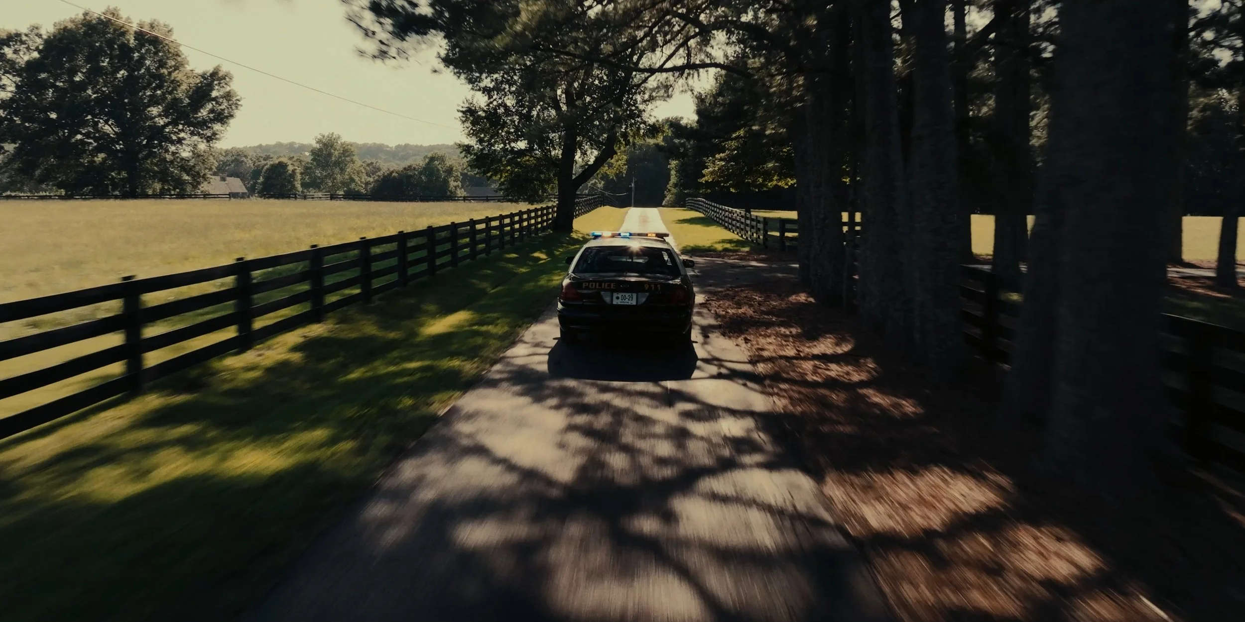 A police car parked on a dirt road beside a wooden fence and trees as the sun sets, casting shadows on the ground.