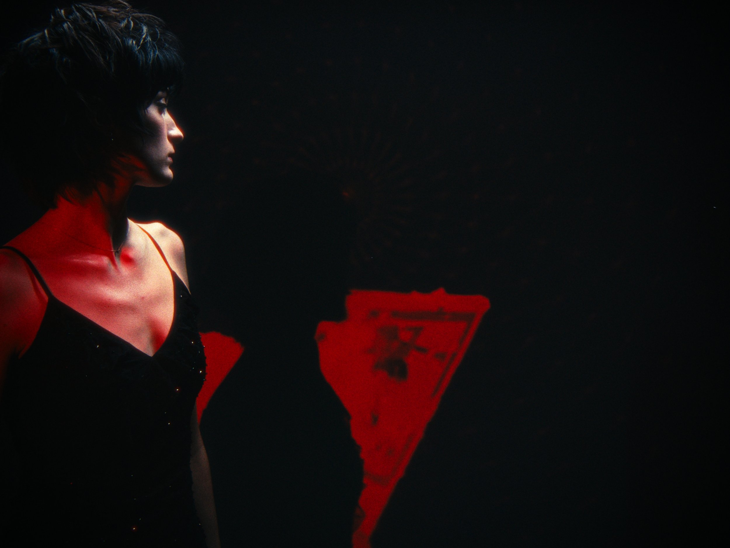 Side profile of a woman with black hair wearing a black dress, standing in dim light with a red object in the background.