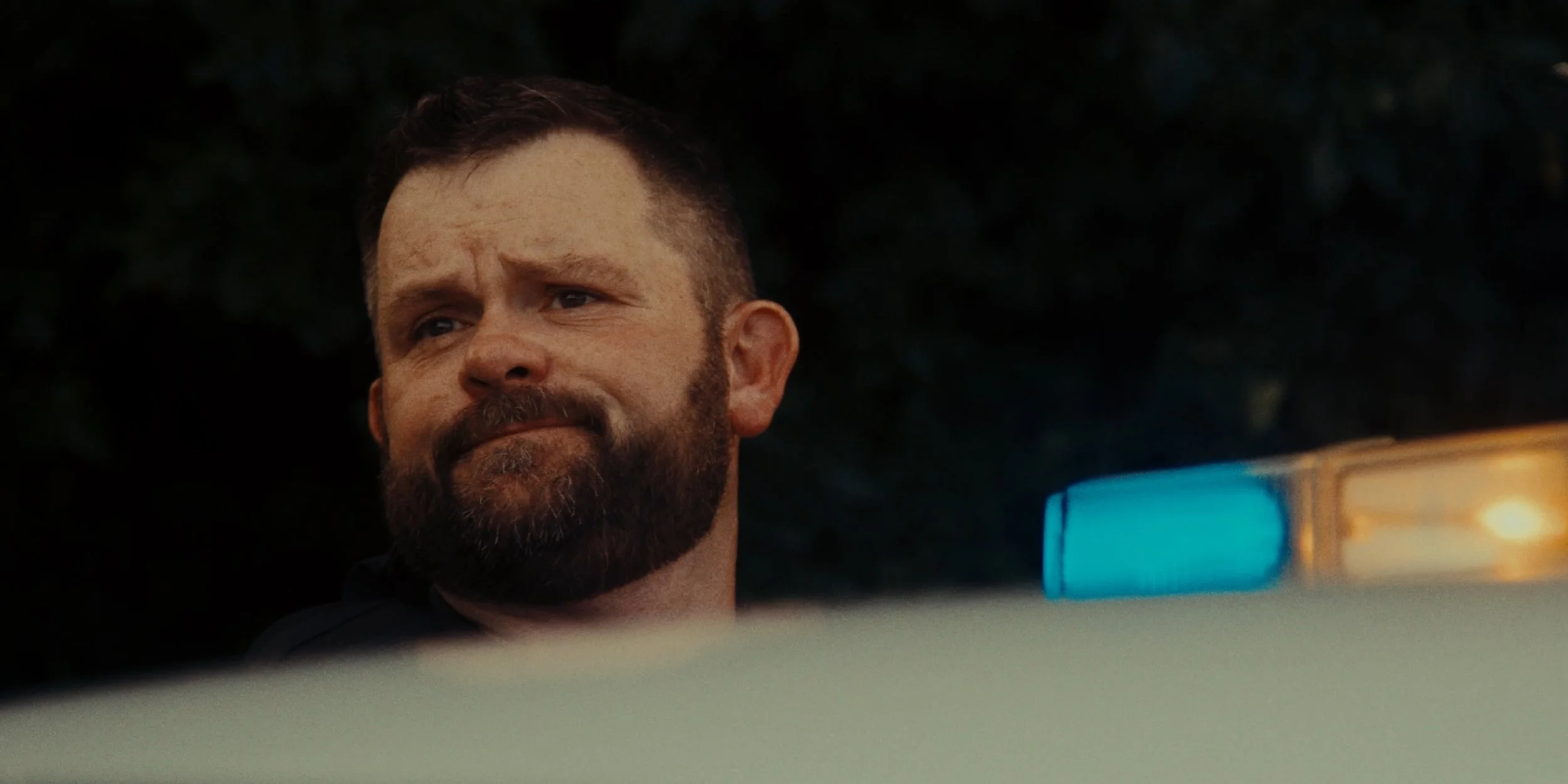 A man with a beard and mustache looks to the side, partially obscured by a vehicle with a blue and orange light bar.