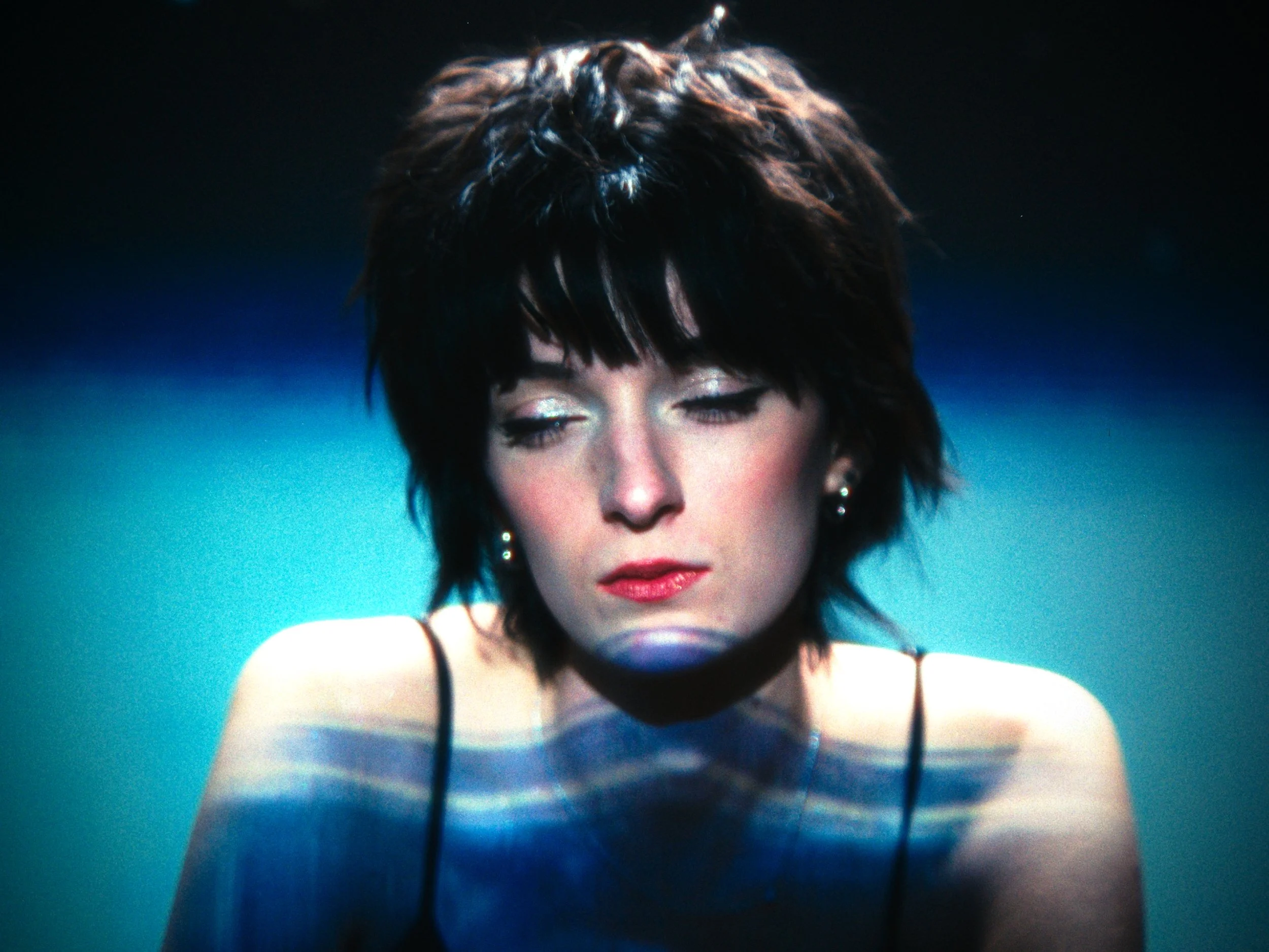 A woman with short dark hair and makeup, wearing a black spaghetti strap top, with a blue gradient background and light patterns projected onto her skin.