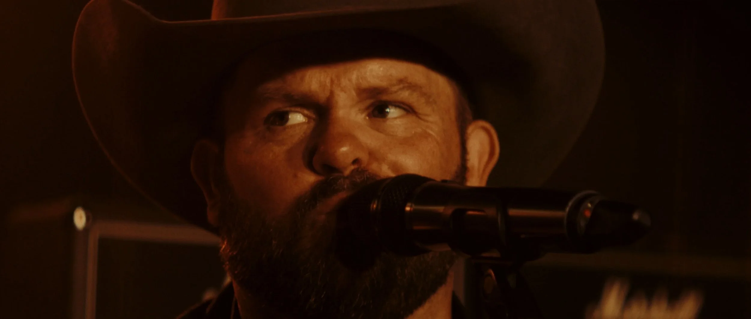 A man wearing a cowboy hat speaking into a microphone in a dimly lit setting.