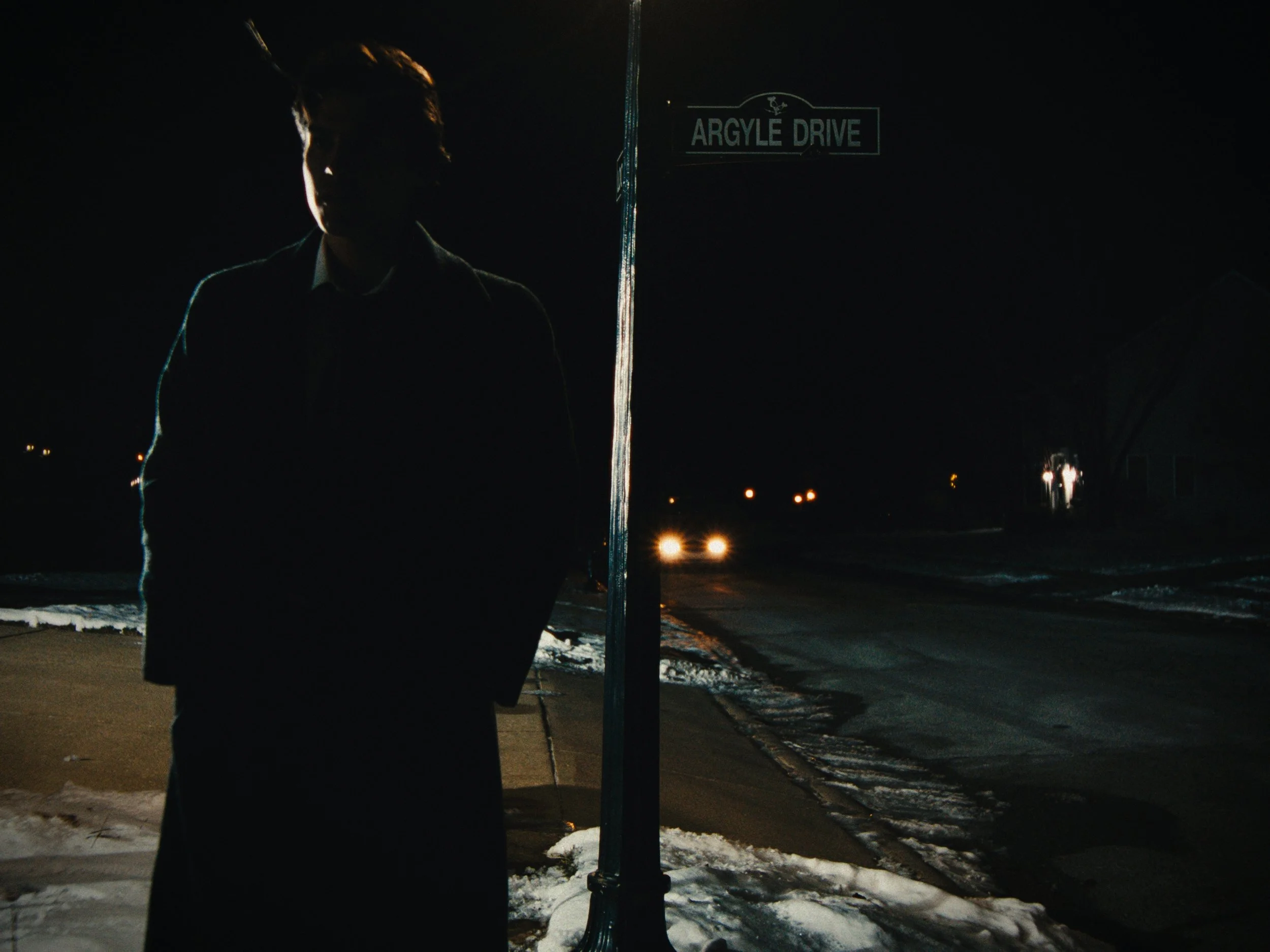 A person standing near a street sign that reads "Argyle Drive" at night. The person is in shadow, with only outline and some hair visible, and there are car headlights approaching in the background. Snow covers parts of the ground.