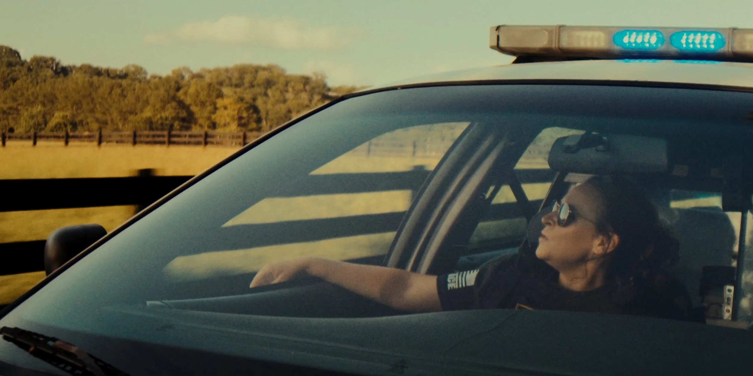 A female police officer wearing sunglasses is sitting in a police car with the lights on, driving along a highway with a wooden fence and trees in the background during the day.