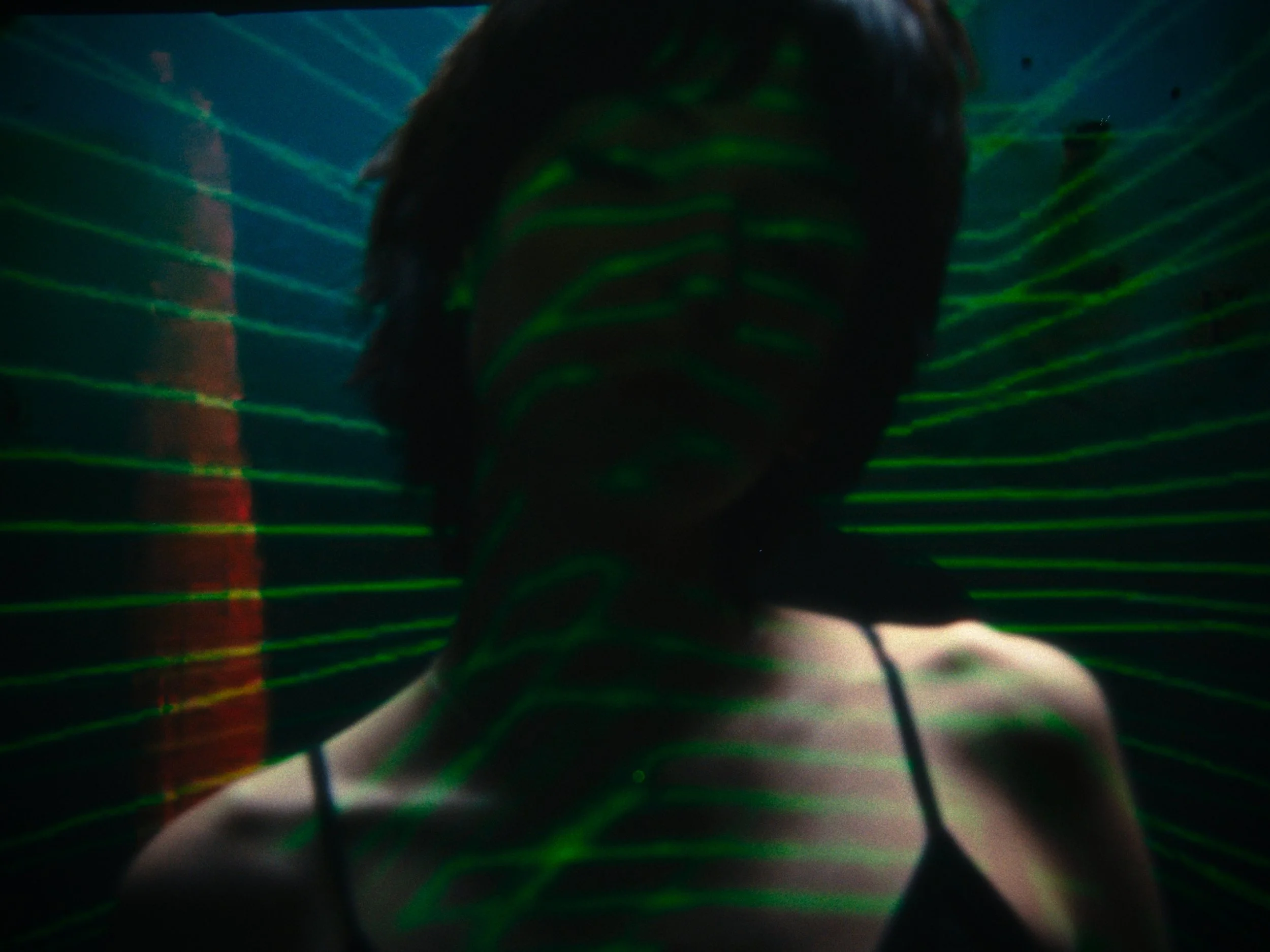 Person with dark hair smiling, wearing a black spaghetti strap top, illuminated by green laser lines creating a pattern on the face and background.