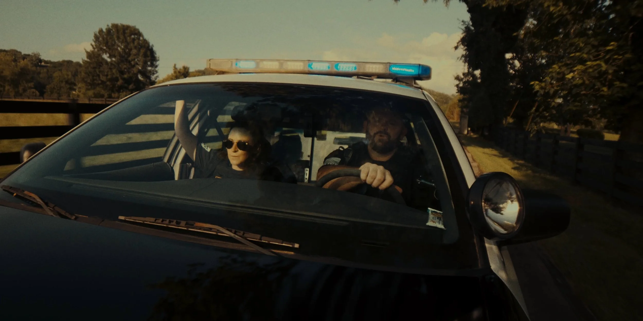 Black and white police car driving on a rural road with two officers inside, one woman wearing sunglasses and a male officer with a beard, surrounded by trees during daytime.