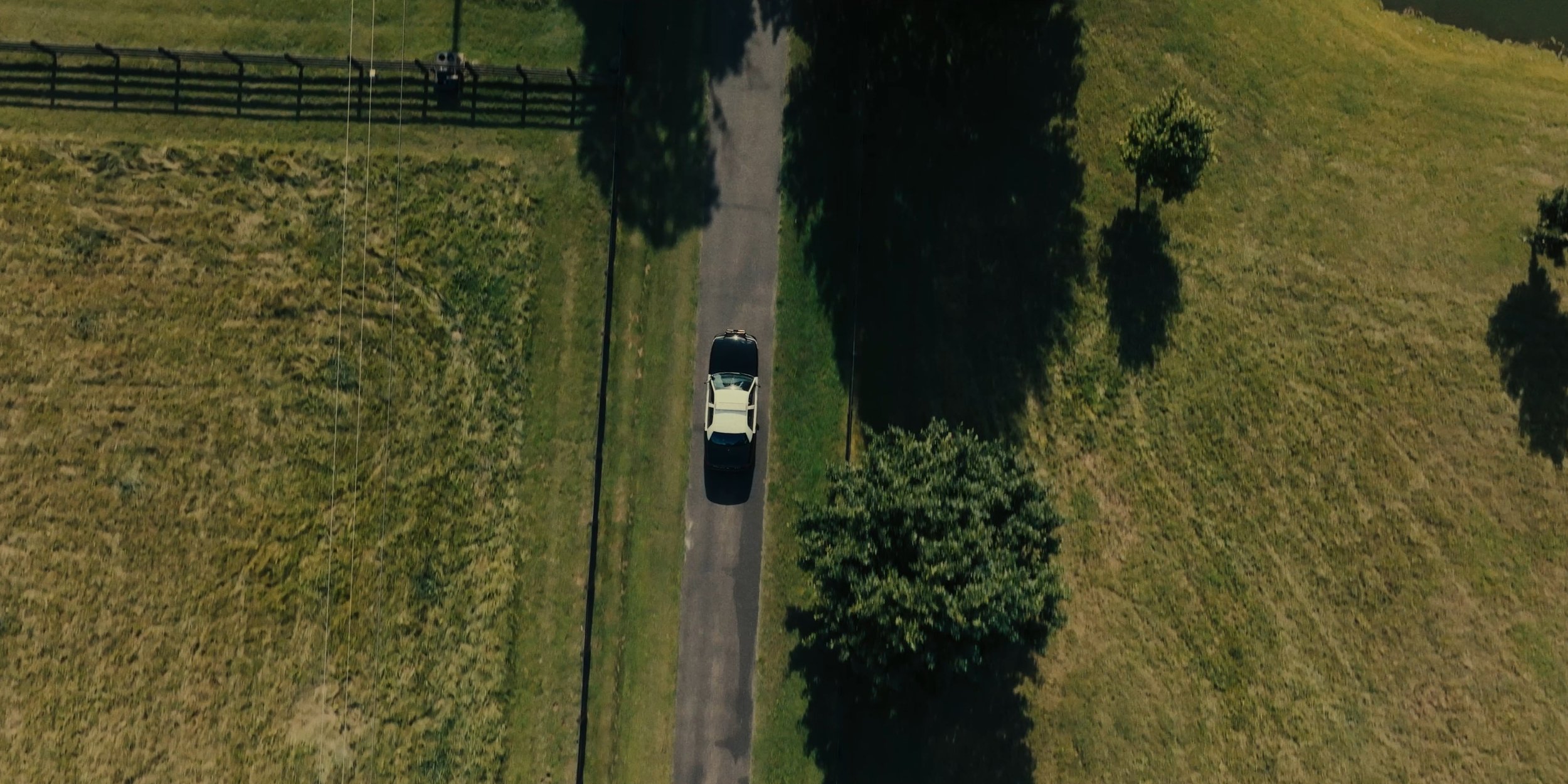 An aerial view of a car driving down a narrow country road surrounded by fields and trees.