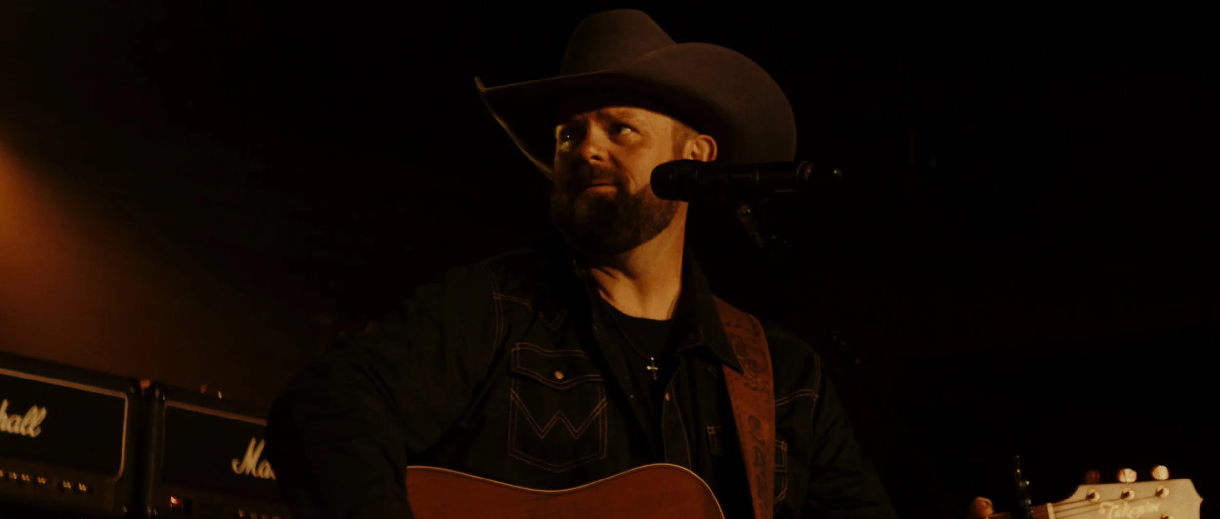 Man wearing cowboy hat playing guitar and singing in a dimly lit setting with Marshall amplifiers in the background.