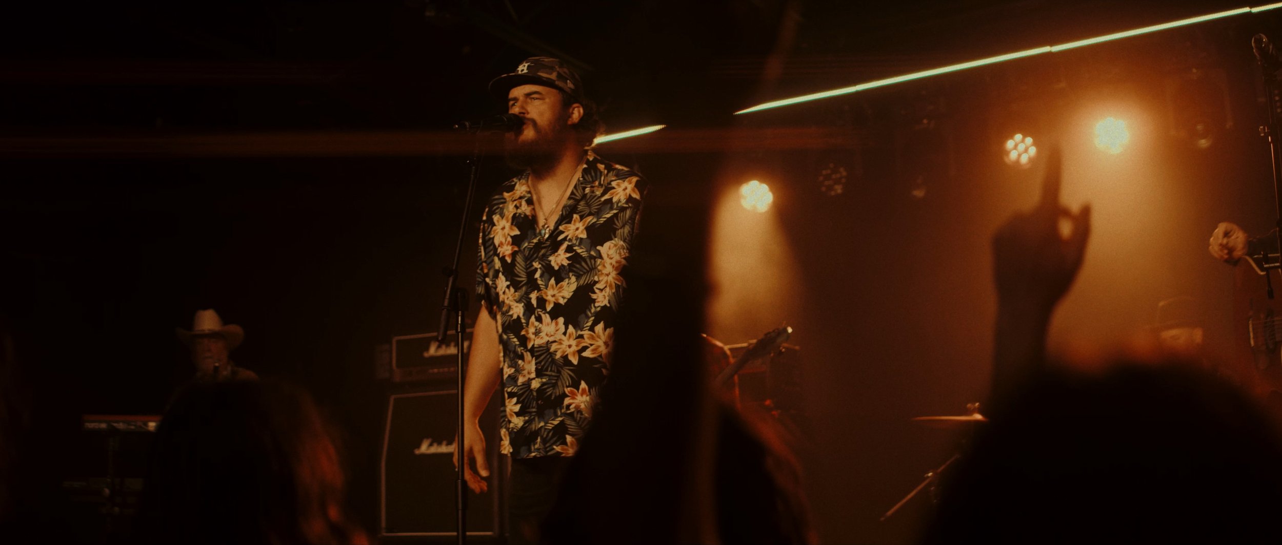 A man singing into a microphone on a stage with warm orange lighting, surrounded by musical equipment and an audience with a raised hand.