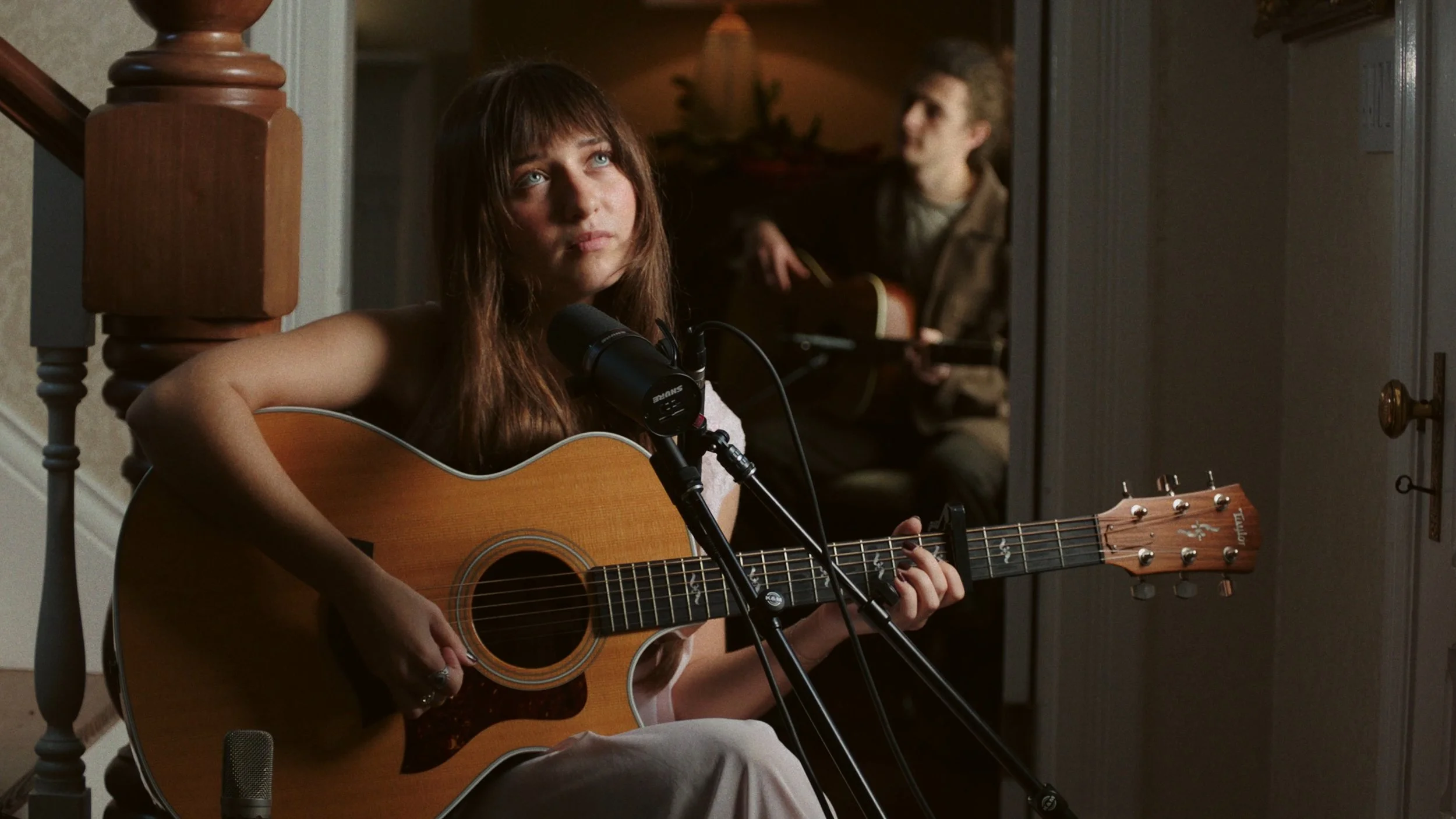 A young woman with brown hair playing an acoustic guitar, singing into a microphone, with a young man playing an acoustic guitar in the background.