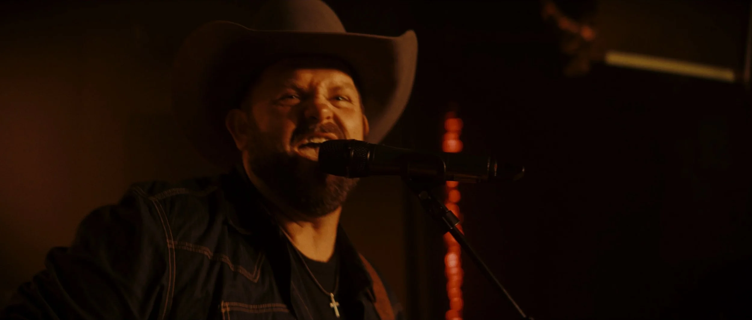 A man singing into a microphone, wearing a cowboy hat and a cross necklace, in a dimly lit setting.