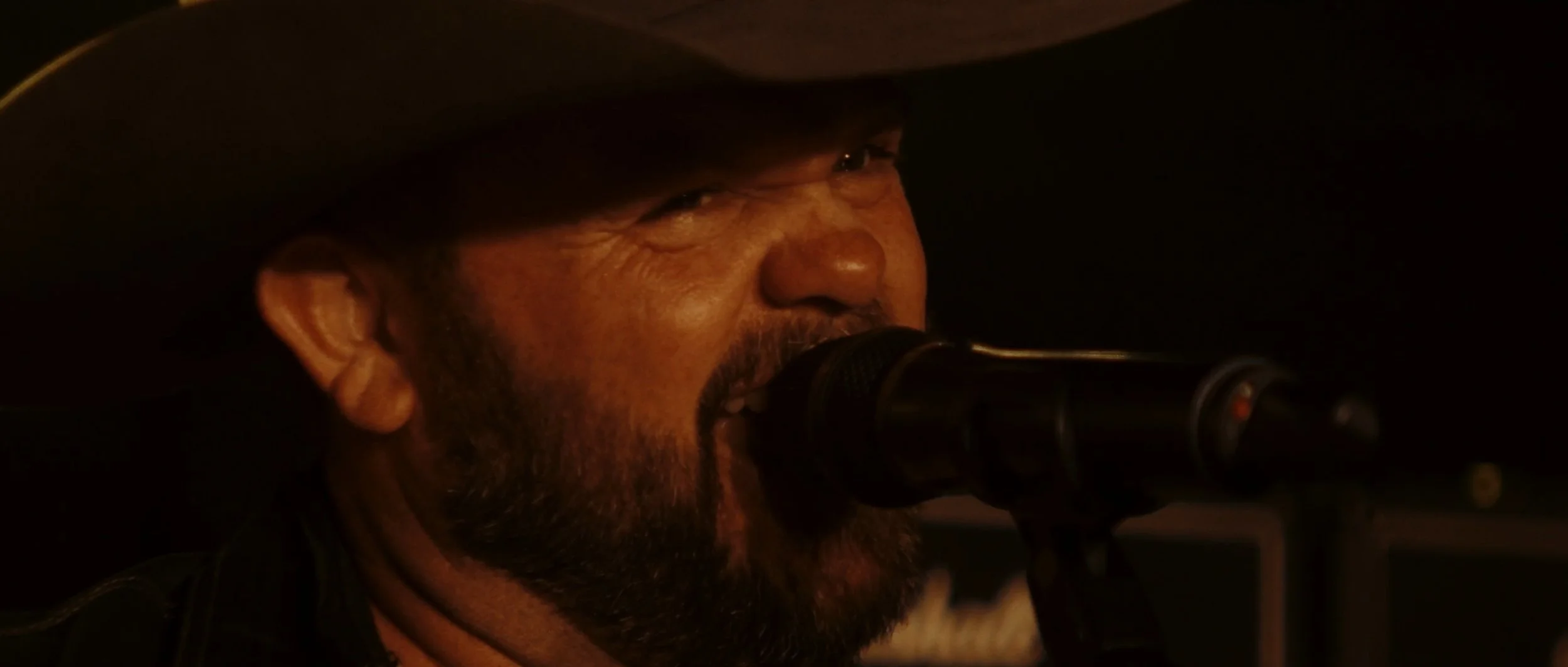 Close-up of a man with a beard wearing a cowboy hat, singing into a microphone in a dimly lit setting.
