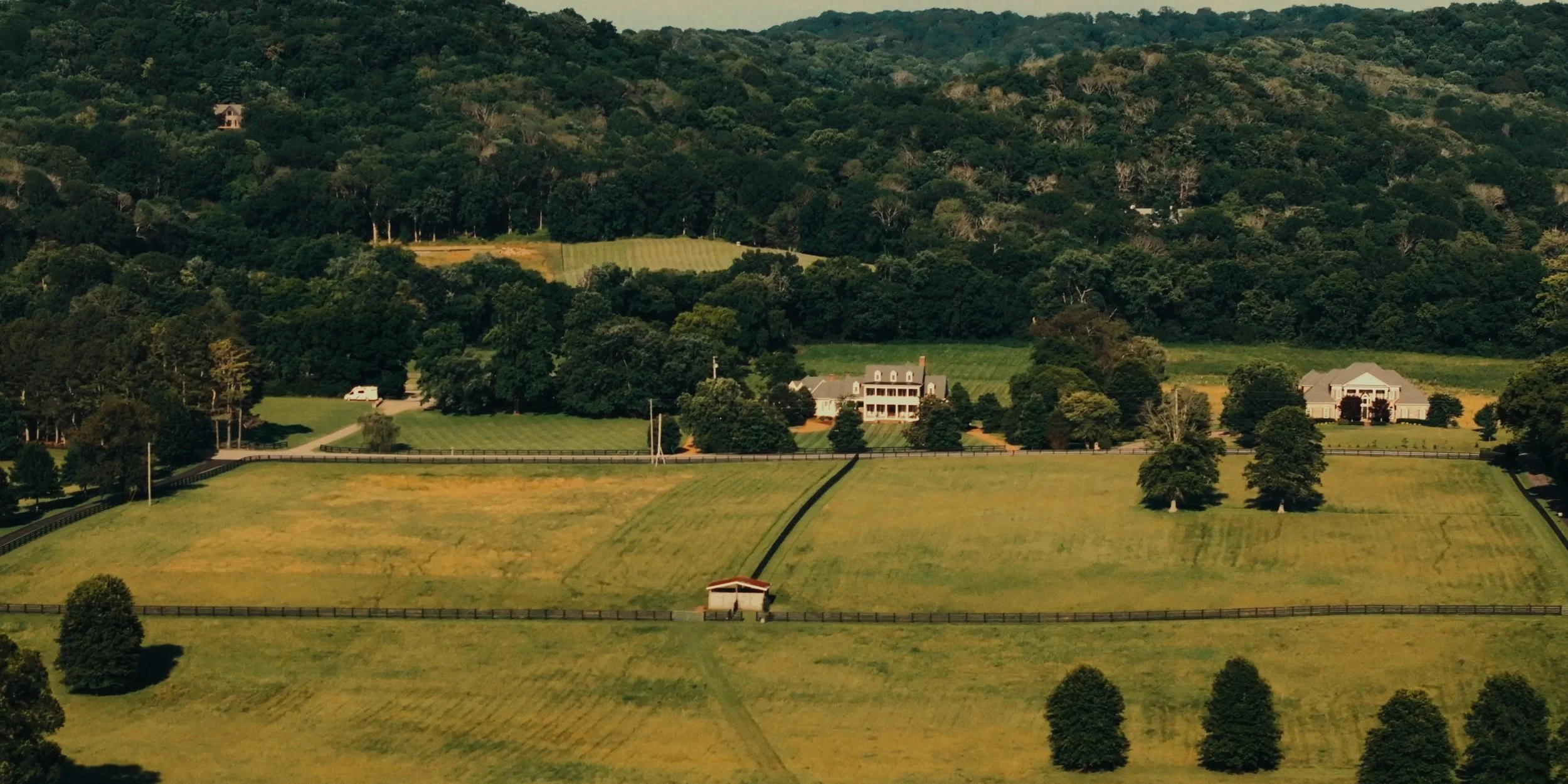 A rural landscape featuring several large houses, green fields divided by fences, and a dense forested hillside in the background.