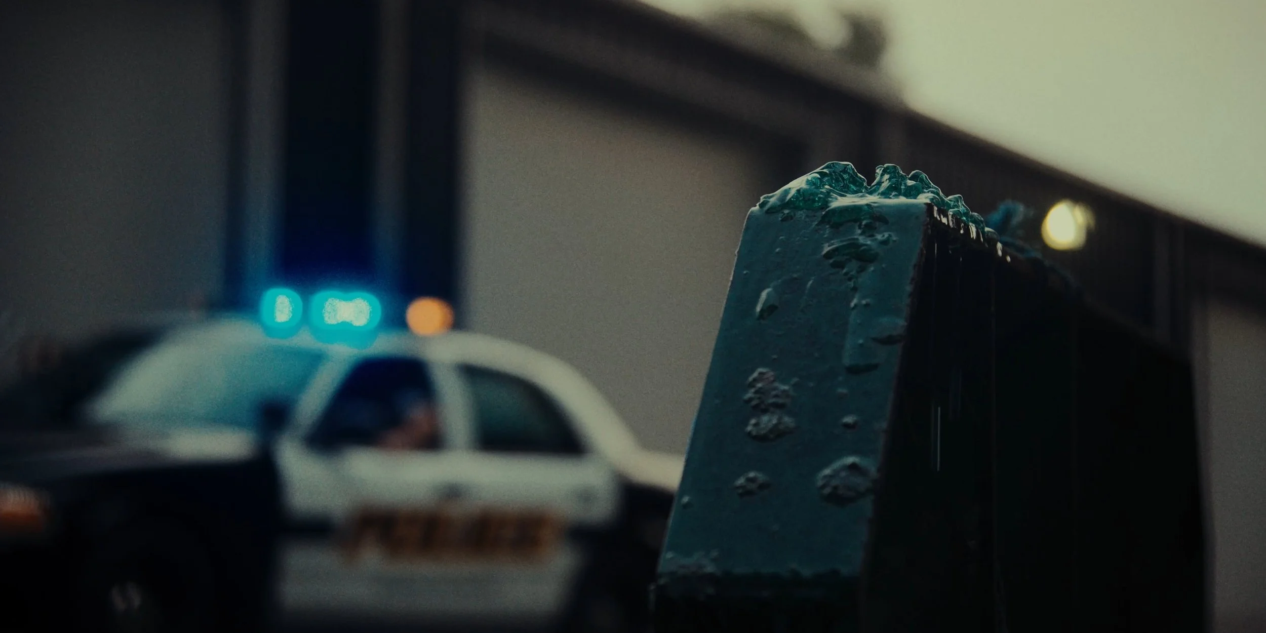 A close-up of a damaged metal surface with rust and debris, with a blurred police car with flashing lights in the background.